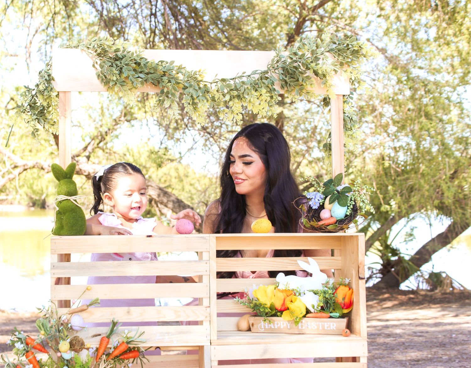 Young mother and baby girl posing together at a spring Easter-themed wooden crate outdoor photo backdrop.