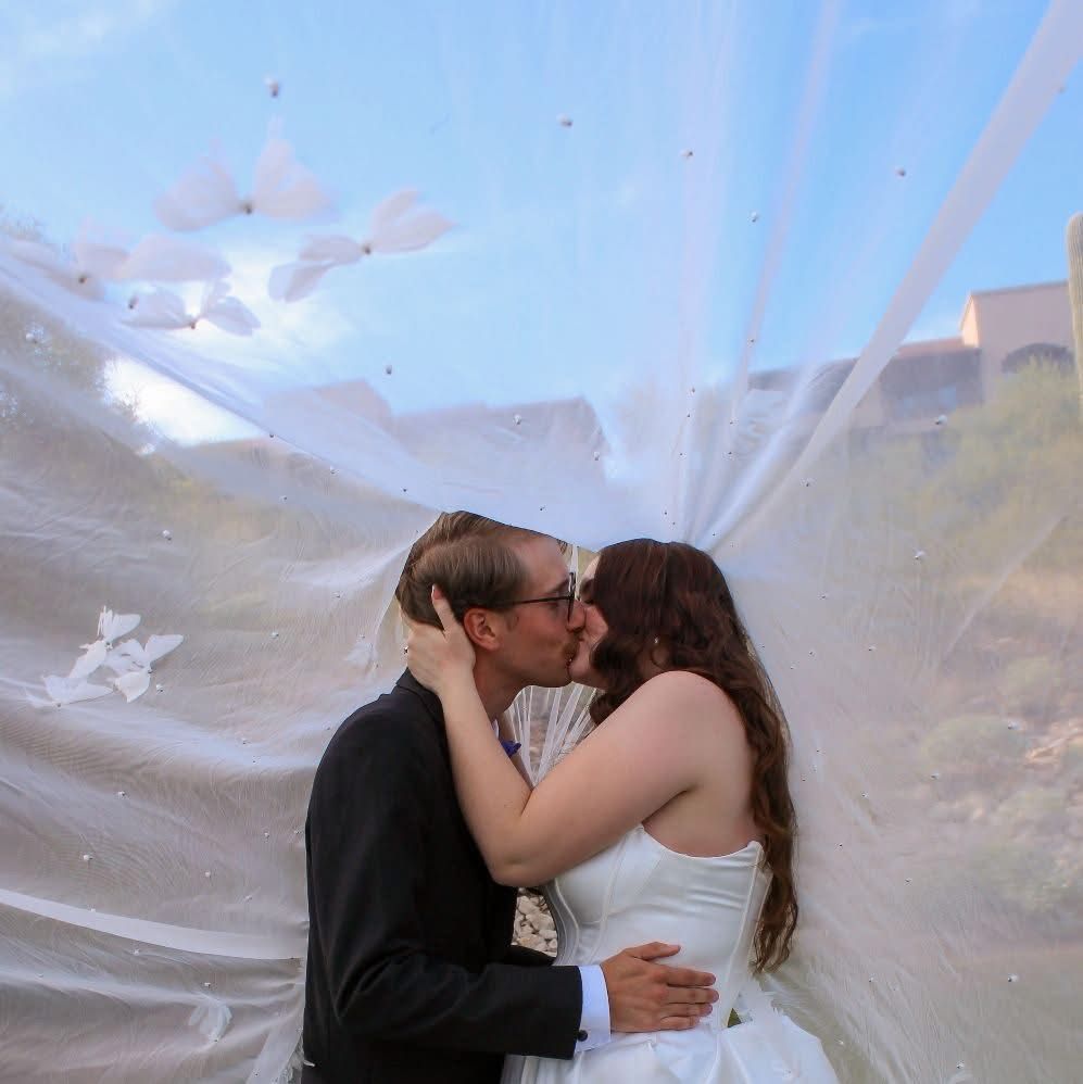 Newlyweds sharing a tender kiss beneath a flowing white veil on their wedding day outdoors.