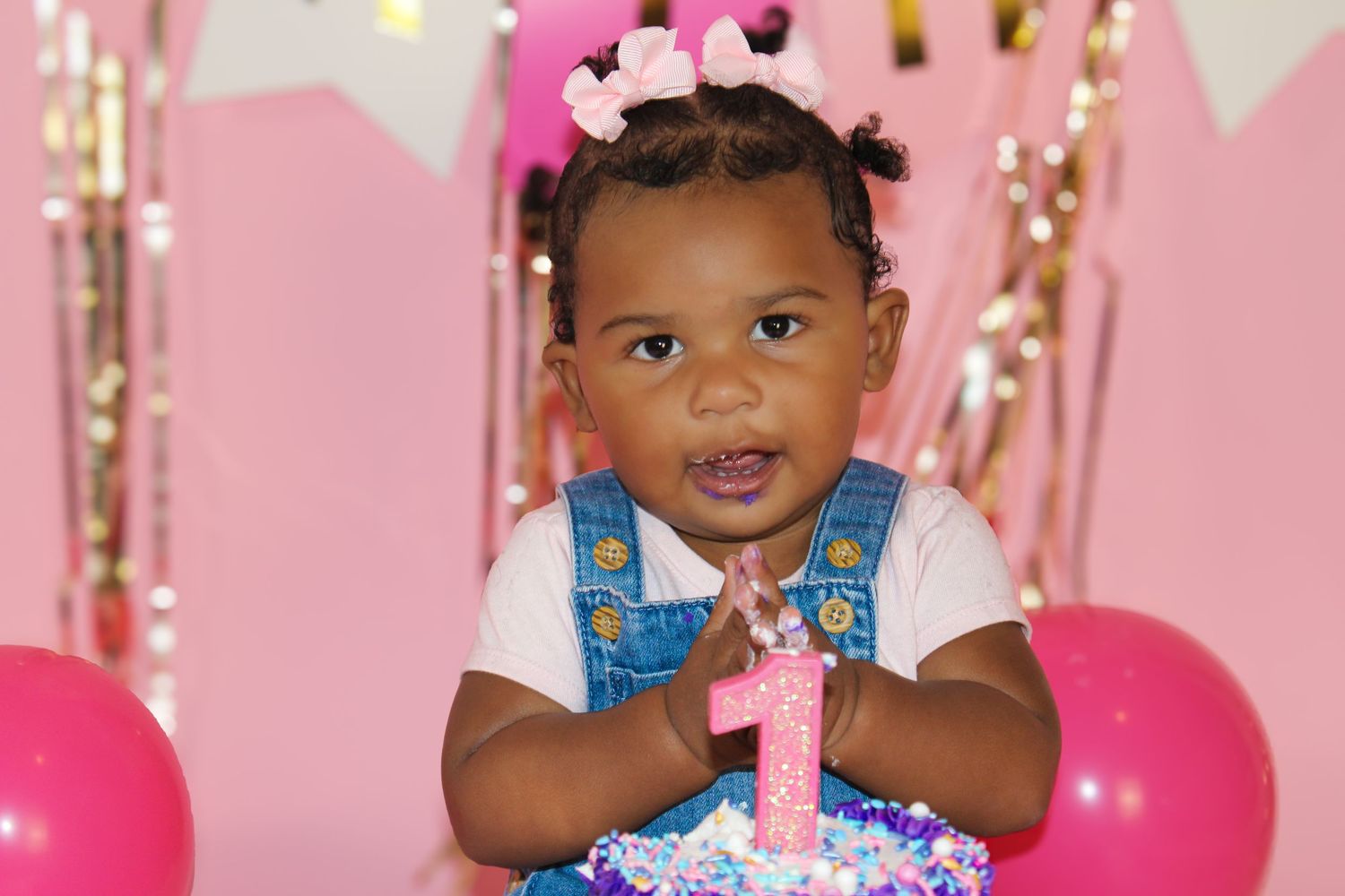 Adorable baby girl celebrating her 1st birthday with a pink cake and balloons in a festive pink setting.