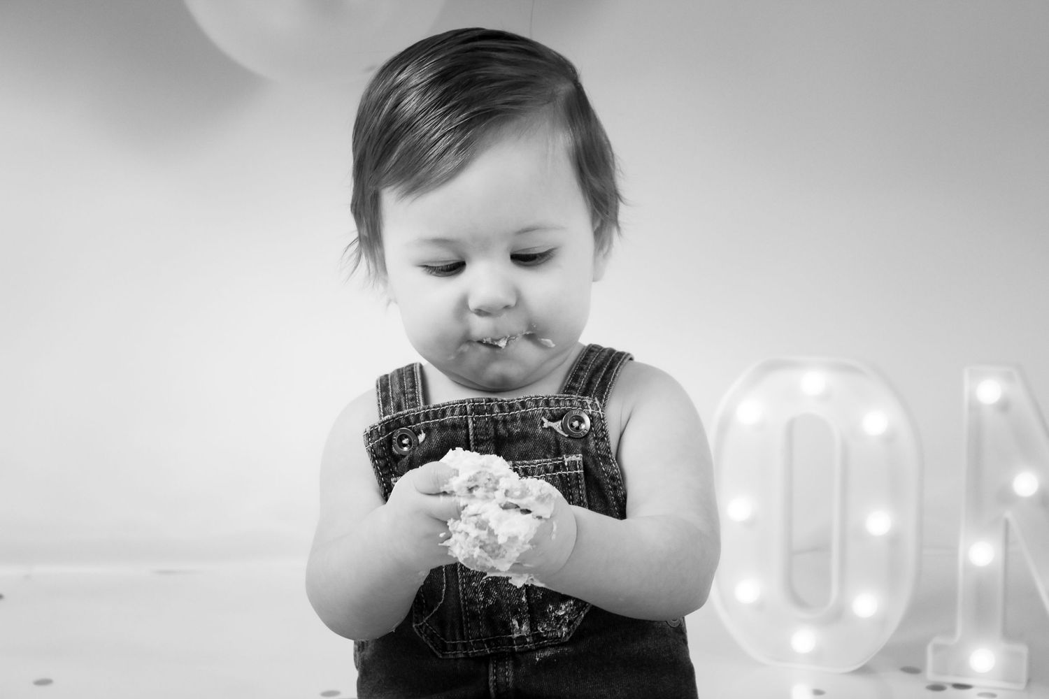 Black and white photo of a baby in overalls eating birthday cake next to a lit marquee letter.