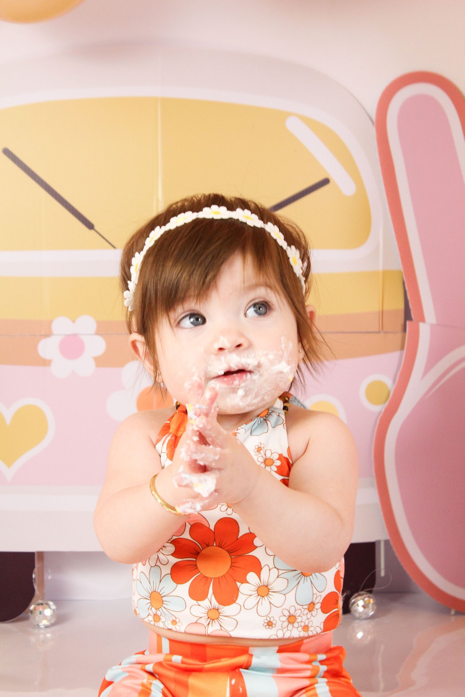 Cute baby girl with cake on her face in a floral dress at a colorful retro-themed birthday party.