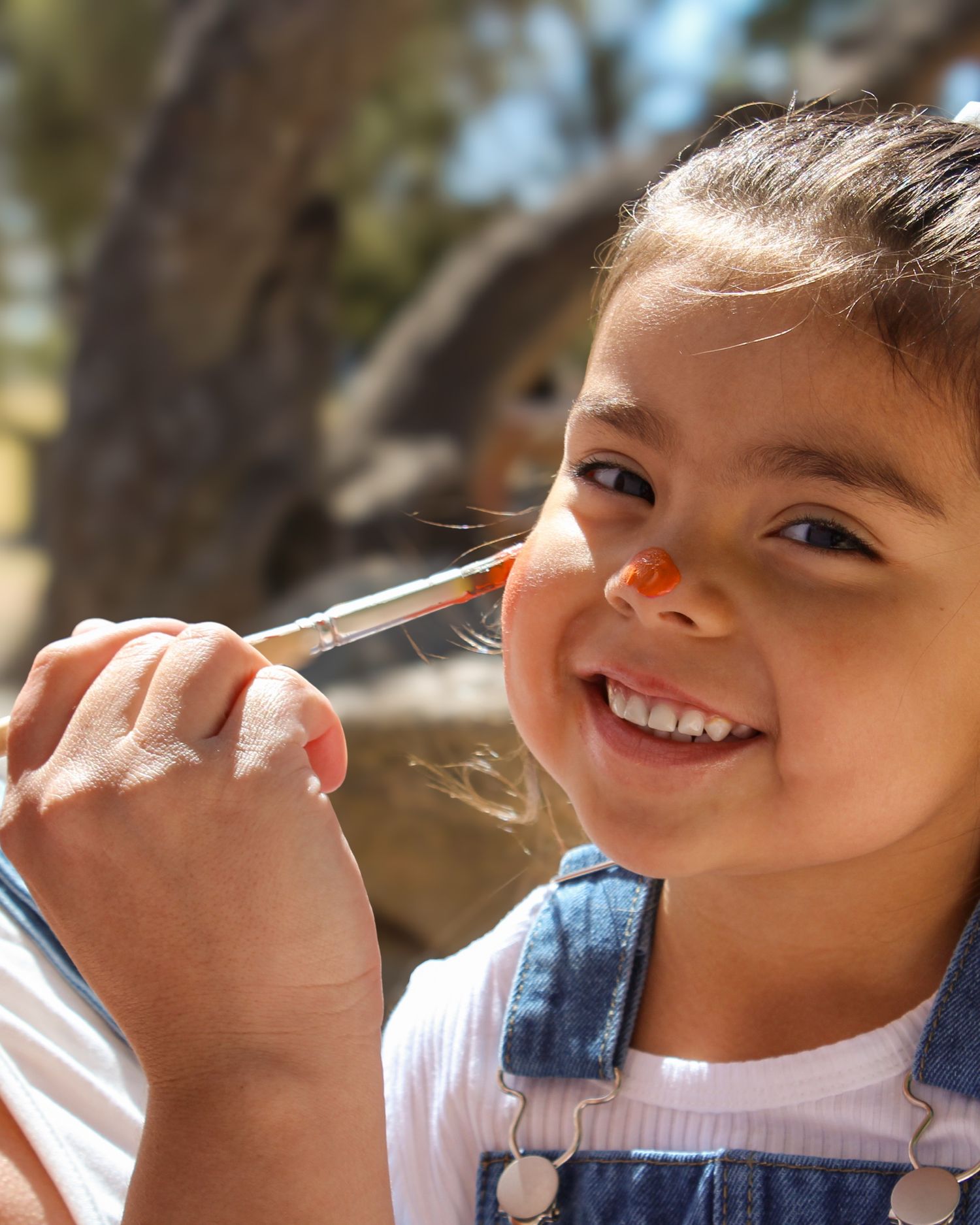 Happy young girl smiling while getting her nose painted orange with a brush outdoors in sunlight.