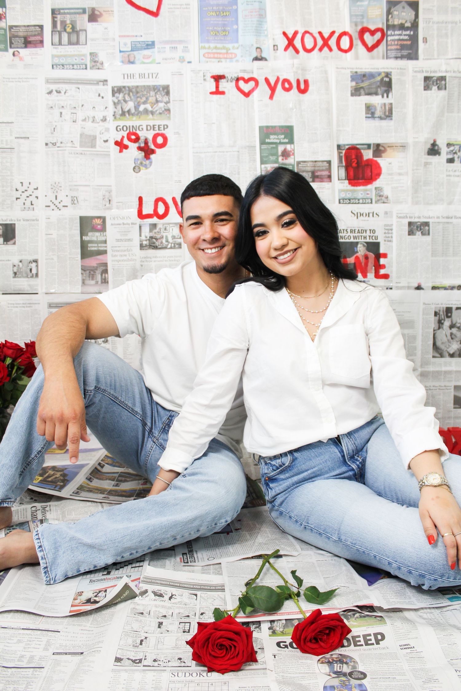 Smiling couple in white shirts and jeans poses with roses on a romantic newspaper Valentine backdrop.