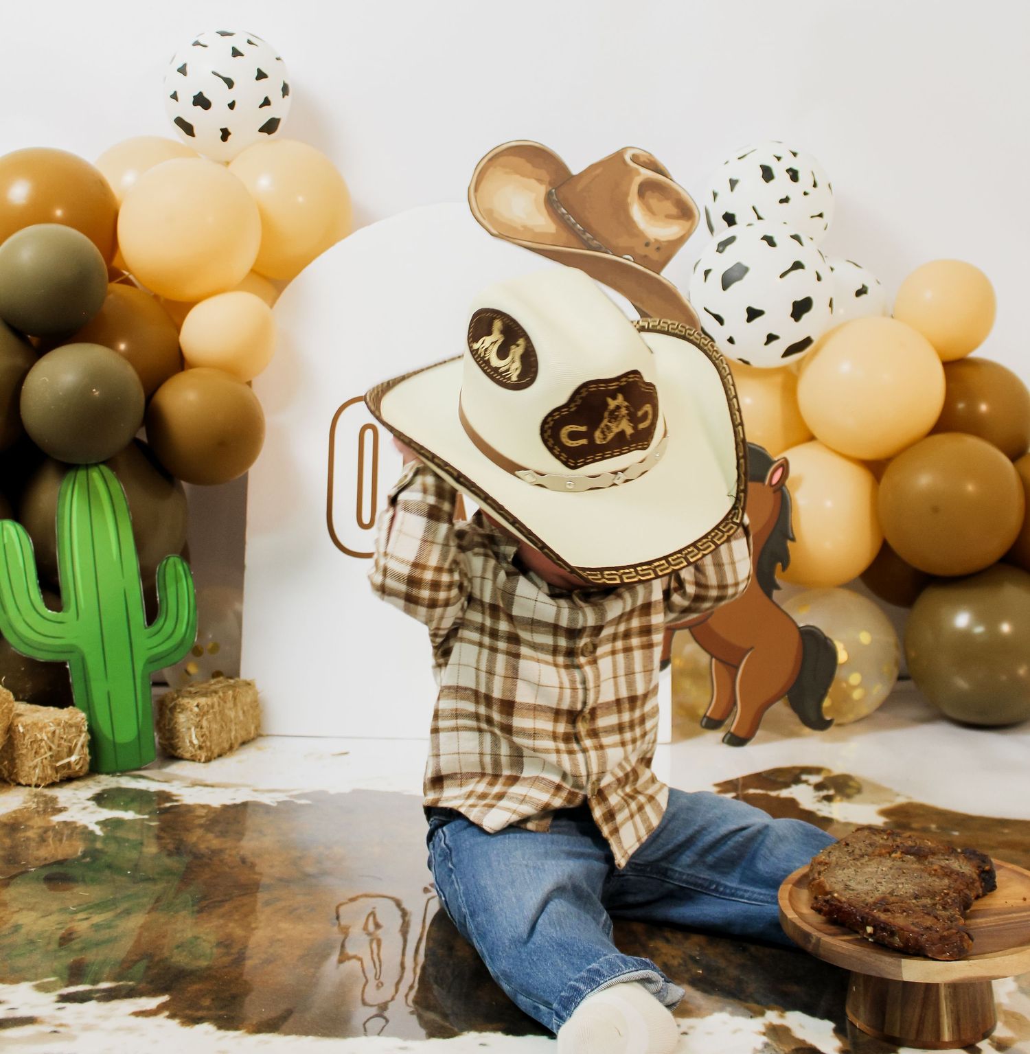 Baby boy in plaid shirt and cowboy hat at western themed first birthday cake smash session.