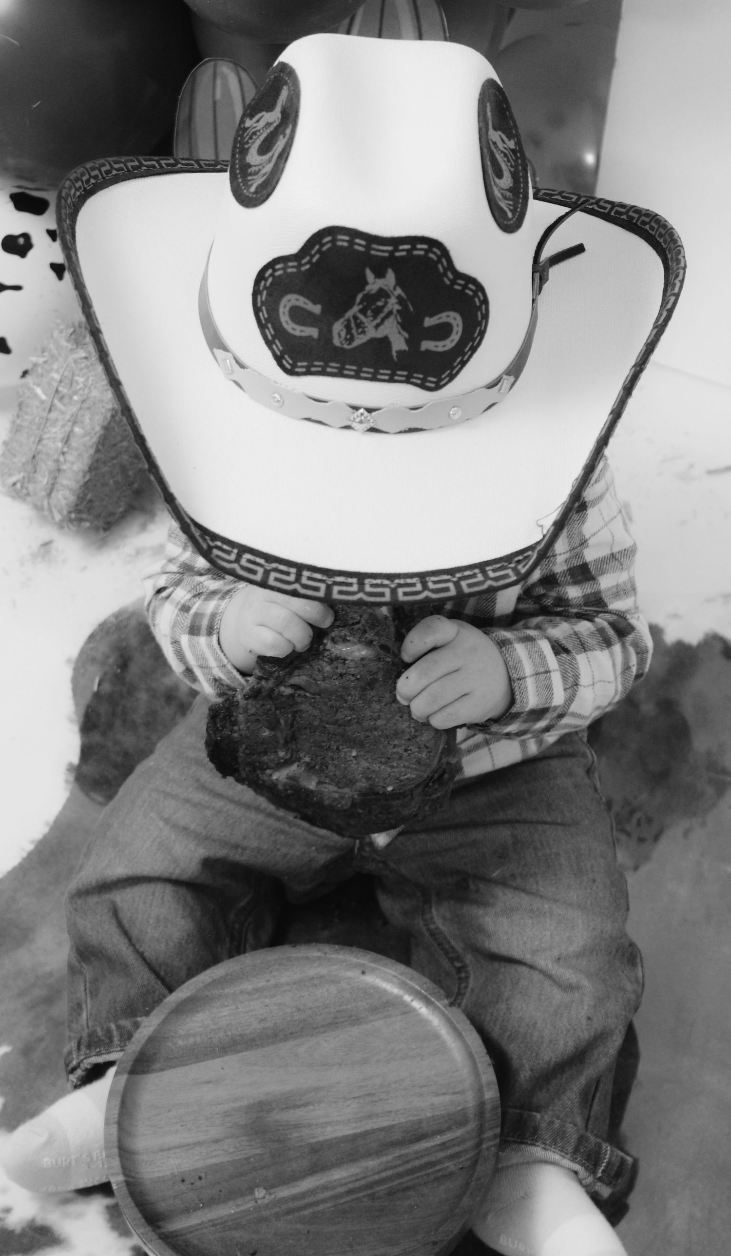 Black and white photo of a baby wearing a cowboy hat and plaid shirt at a western themed party.