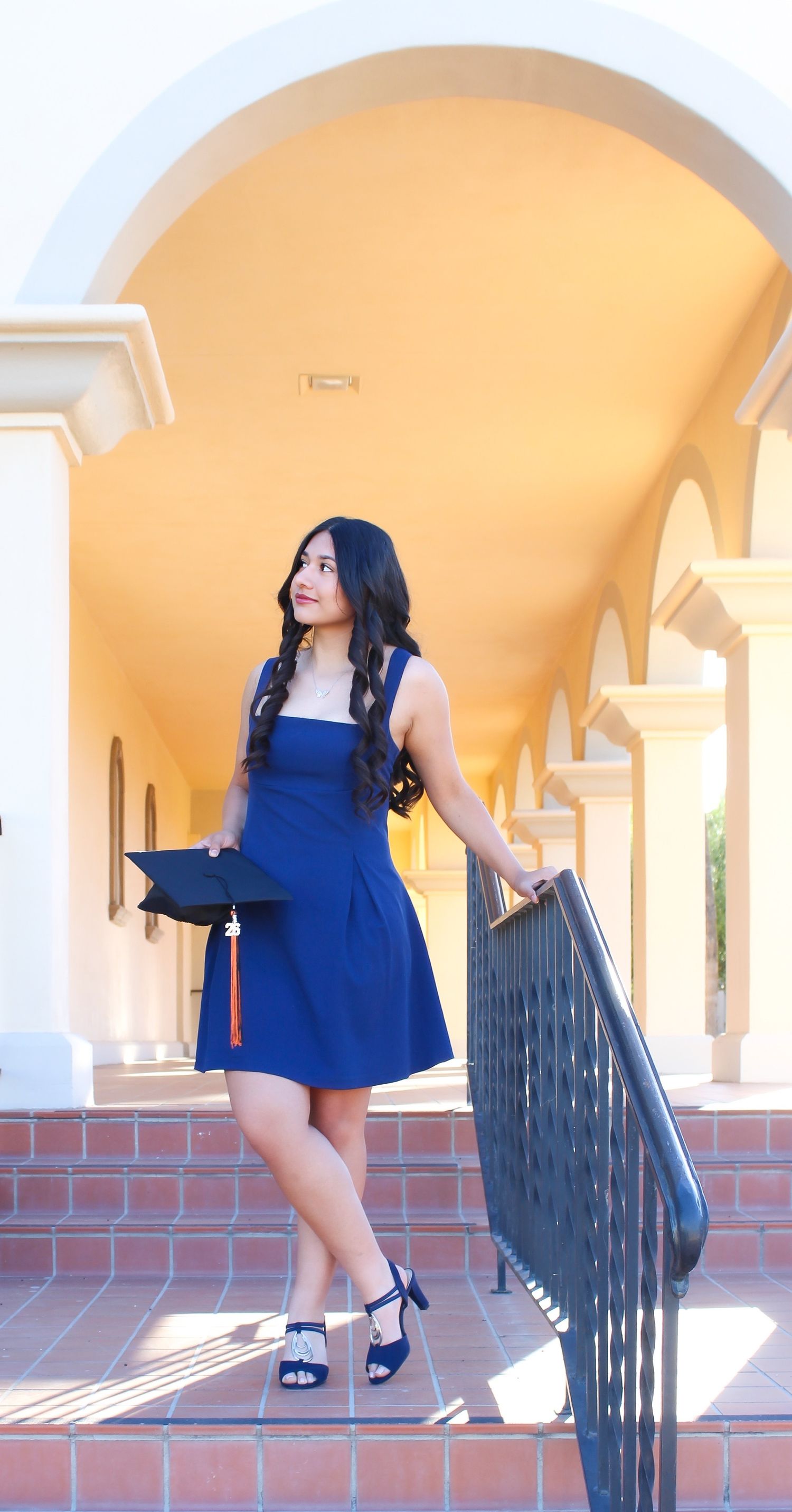 Young woman in blue dress holding graduation cap while standing on steps under an arched building entrance.