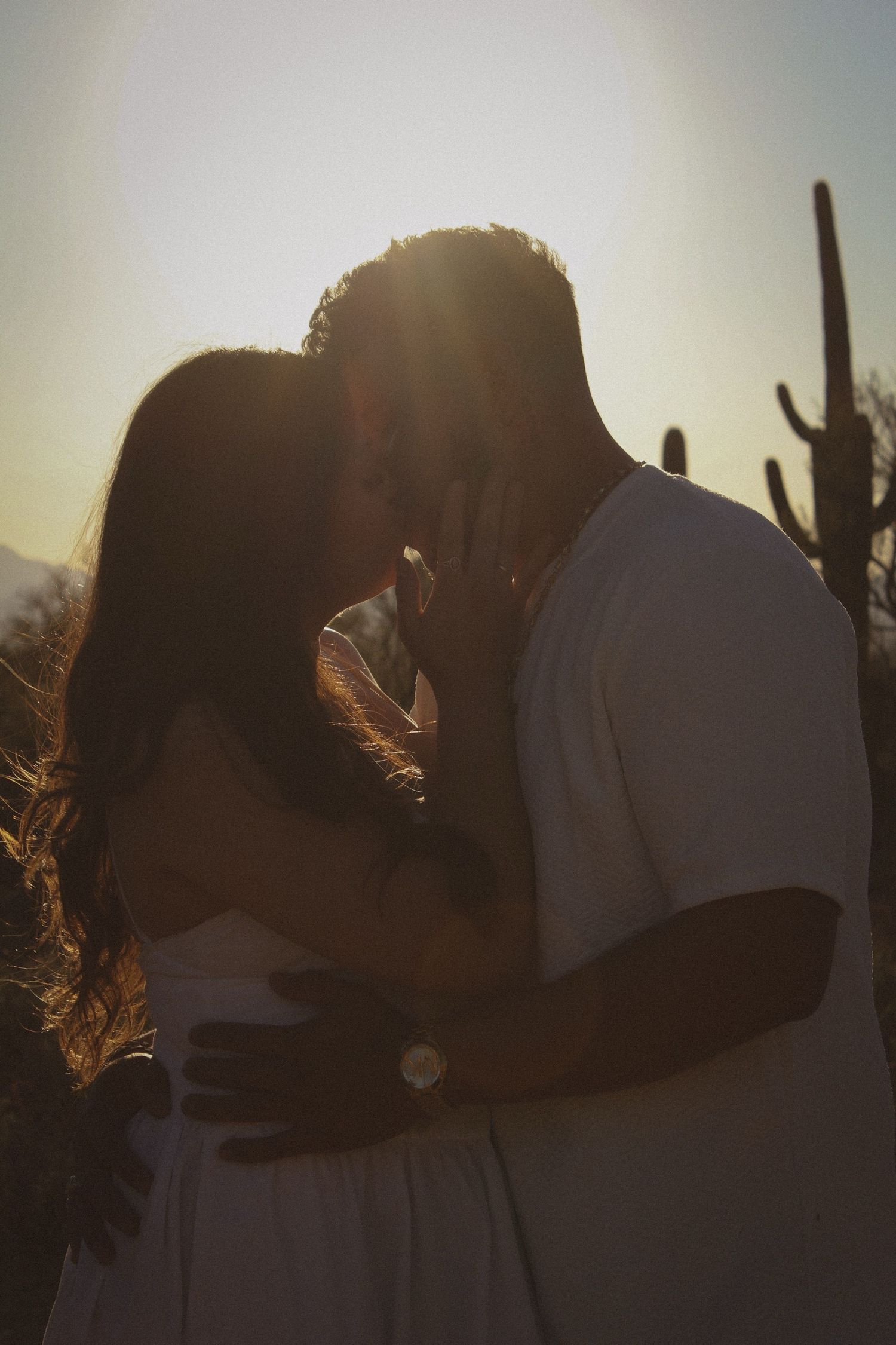 Couple sharing romantic kiss silhouetted against golden sunset with saguaro cactus in background.
