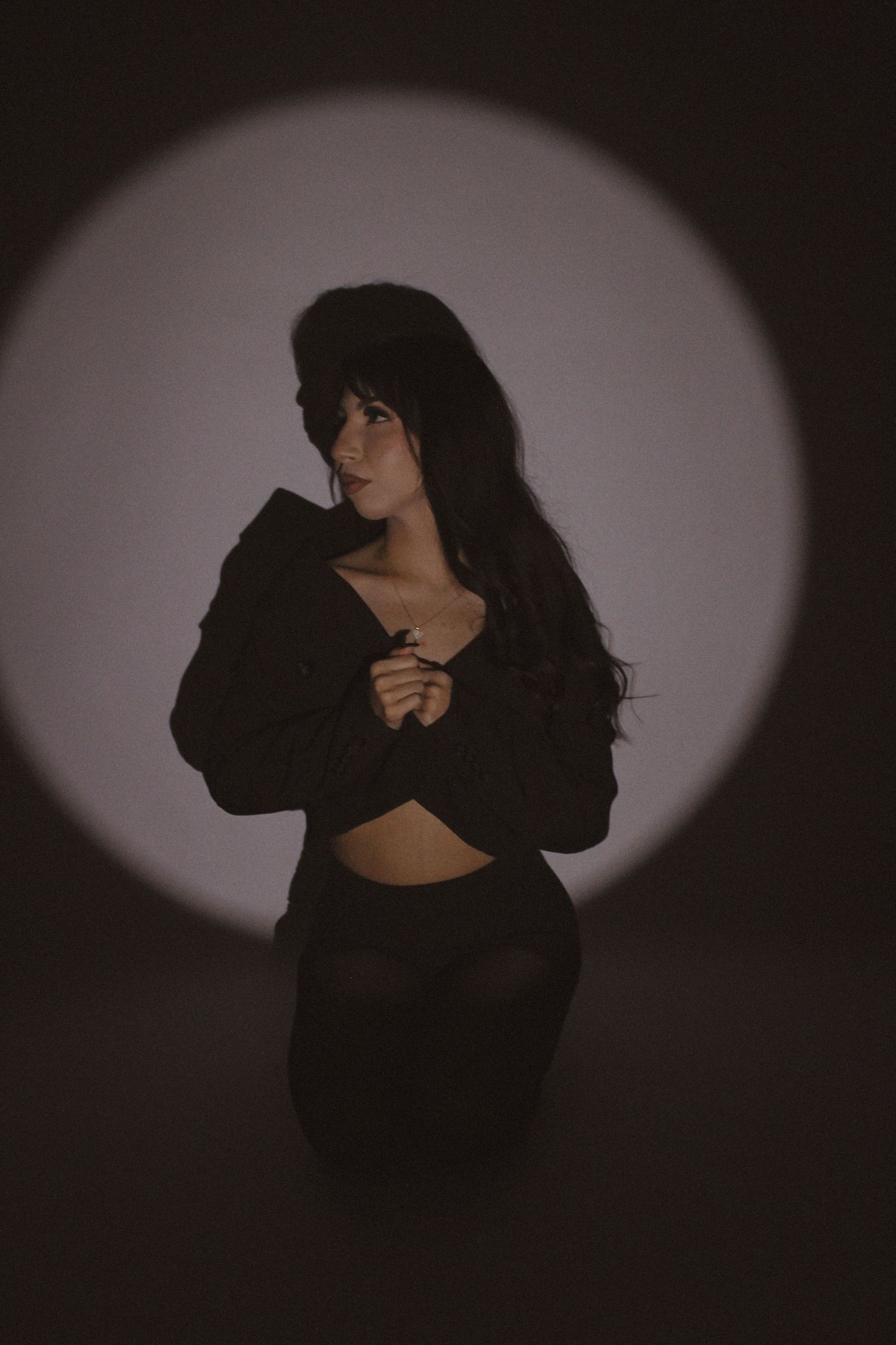 Woman in black crop top and pants posing dramatically in circular spotlight against dark background.