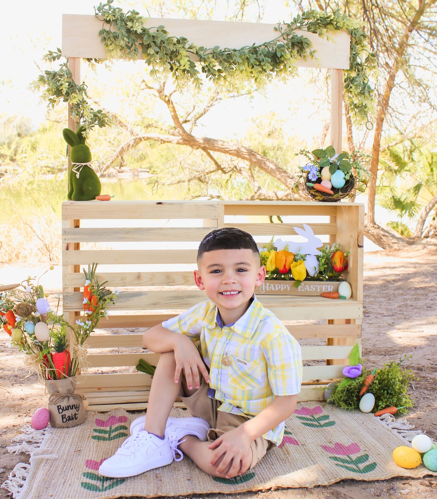Smiling young boy in yellow plaid shirt sitting at a festive Easter outdoor photo backdrop with eggs.