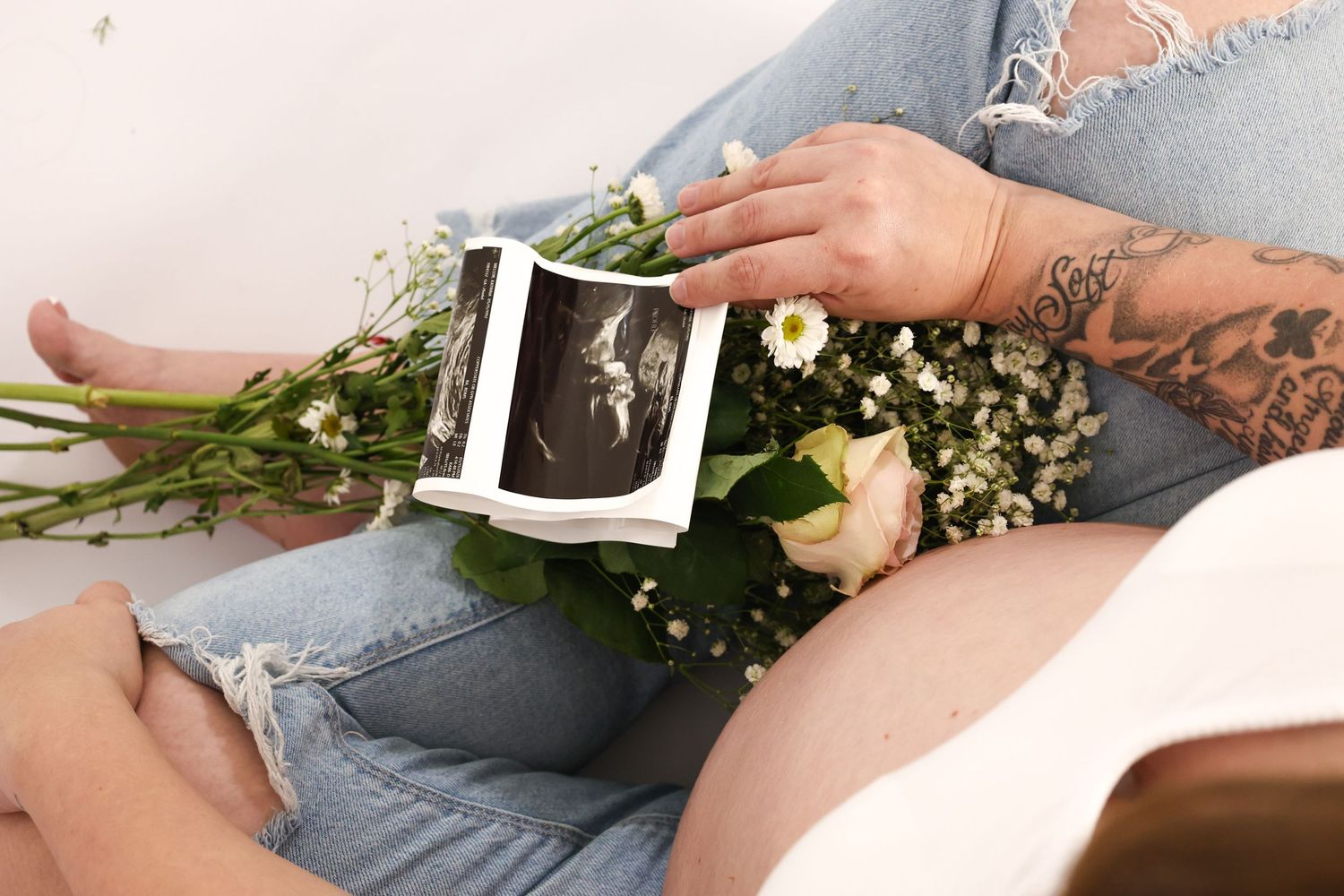 Close-up of tattooed pregnant woman holding ultrasound photos with a floral bouquet in lap.