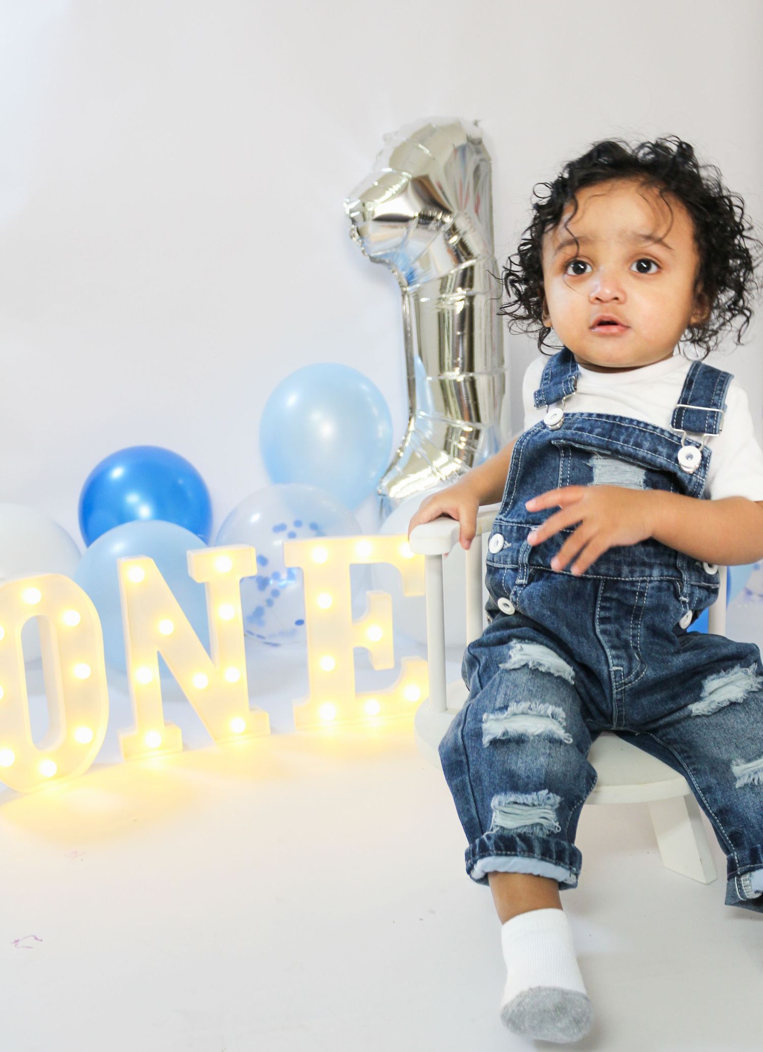 Baby boy in denim overalls posing with ONE marquee letters and blue balloons for first birthday.