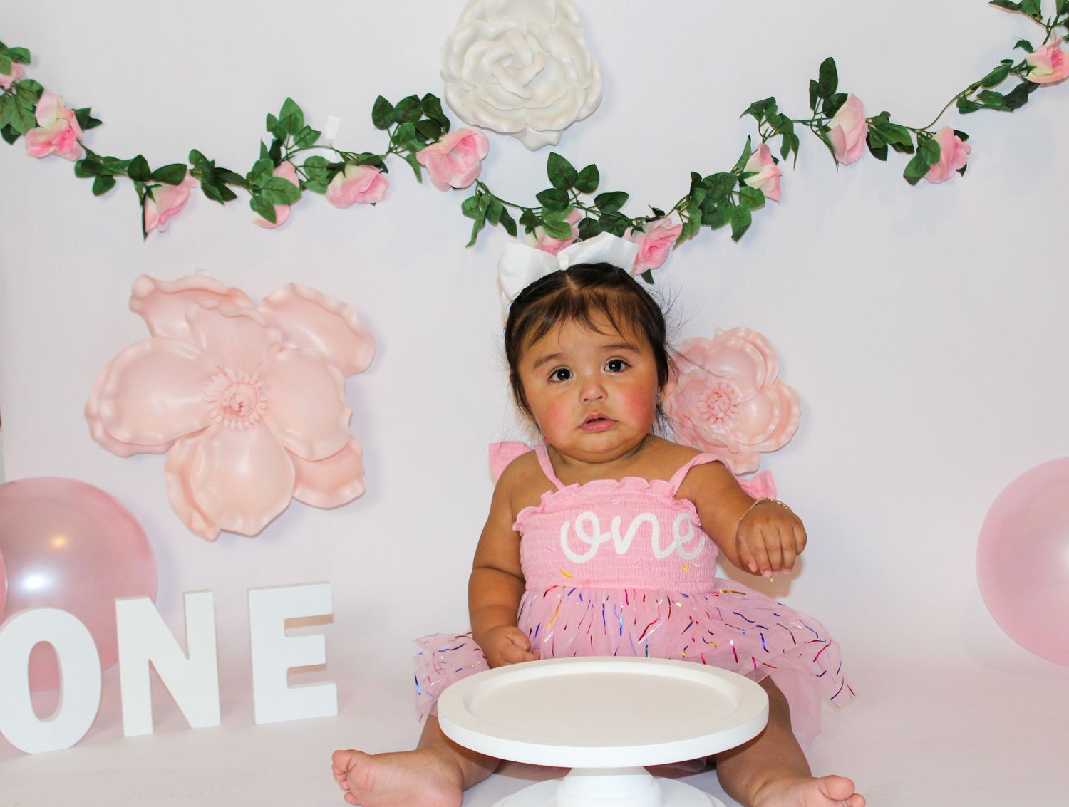 Baby girl in pink tutu sitting with white cake at floral first birthday cake smash session.