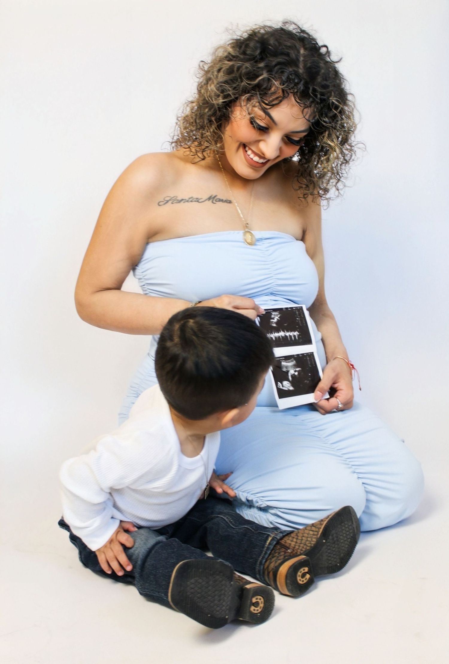 Pregnant woman in blue tube dress smiling at ultrasound photos held by her young curly-haired son.