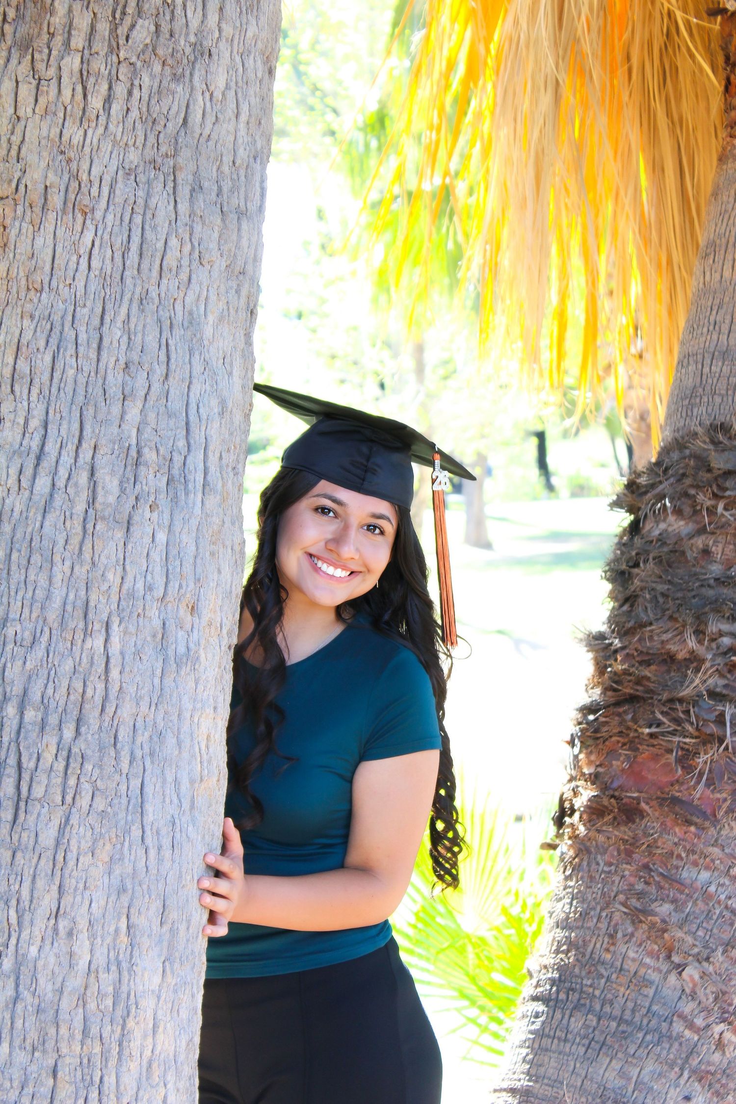 Happy young female graduate wearing a black mortarboard cap smiling between two palm trees outdoors.