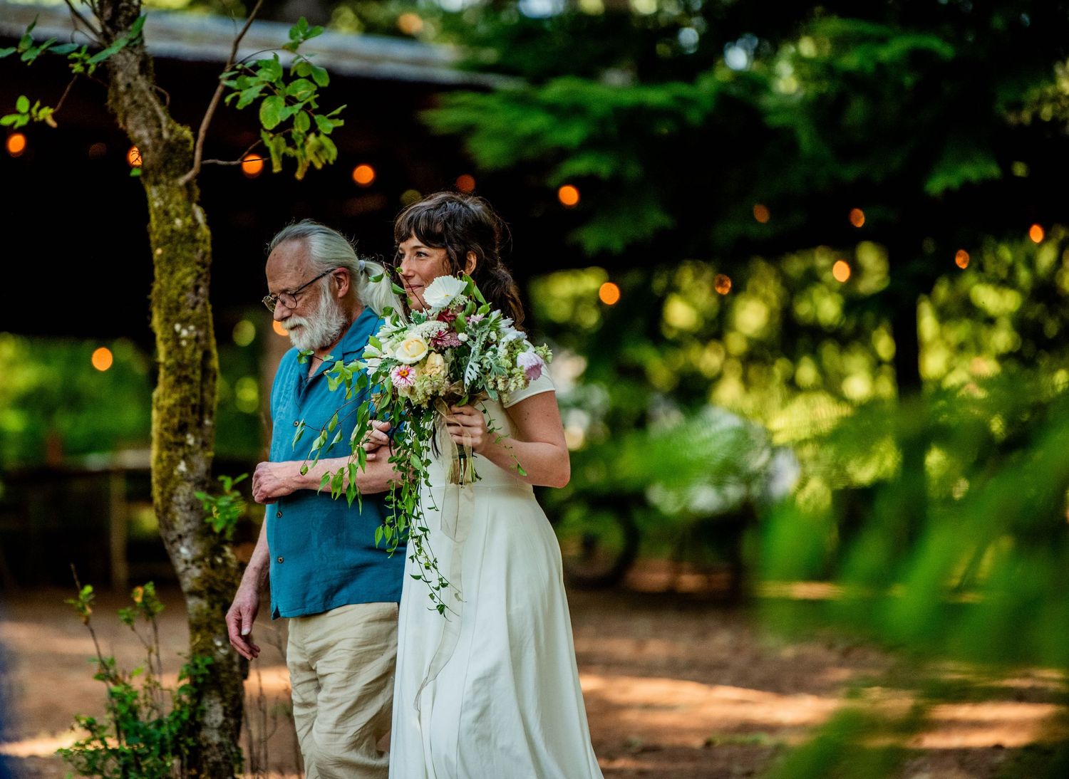 A proud father walks his daughter down the aisle in a verdantly green outdoor summer wedding.