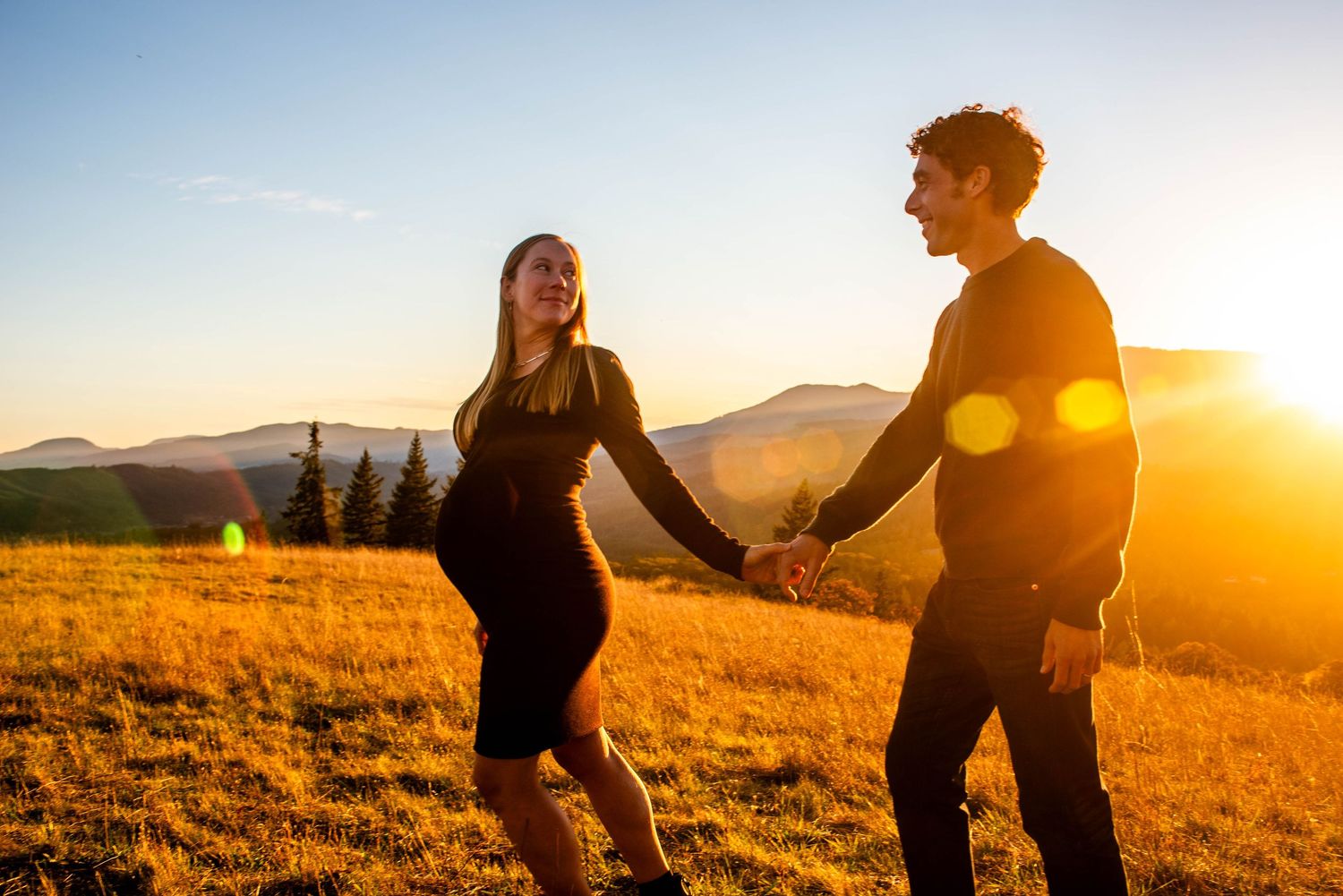 An adorable couple with a baby on the way hold hands and gaze at each other as they walk through golden evening light with mountains behind.
