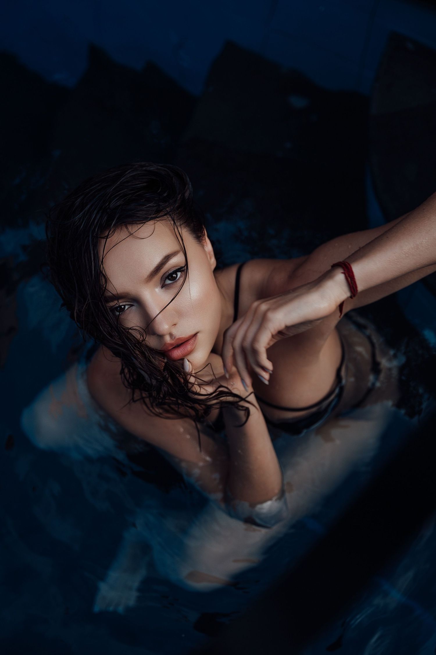 Young woman with wet hair emerging from dark pool water, wearing a black swimsuit and red bracelet.