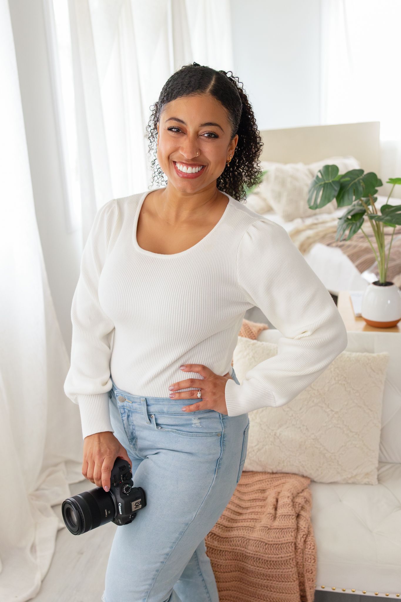 Professional photographer in white blouse and light jeans poses with camera in bright modern studio setting.
