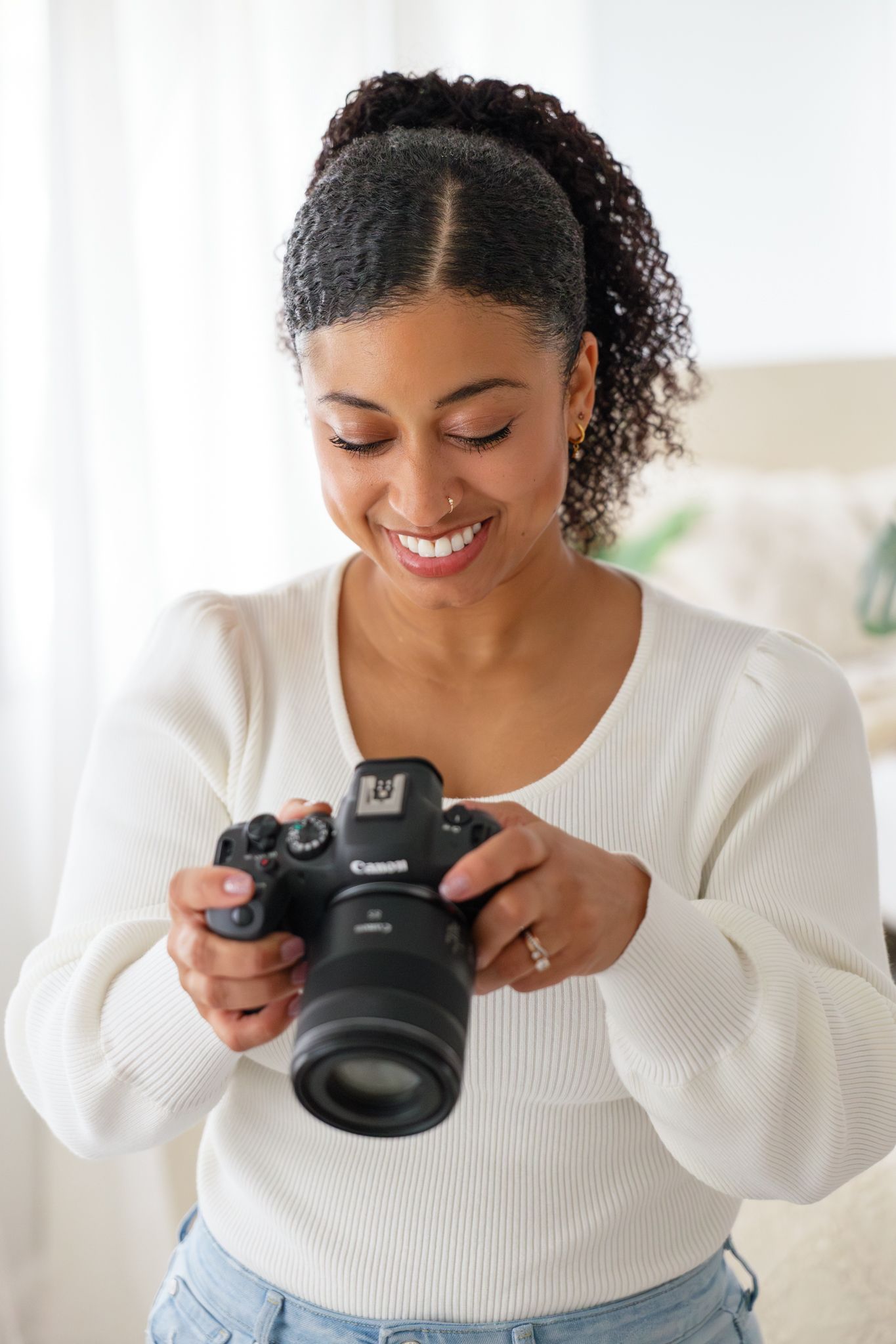Photographer in white sweater smiling while looking down at professional DSLR camera.