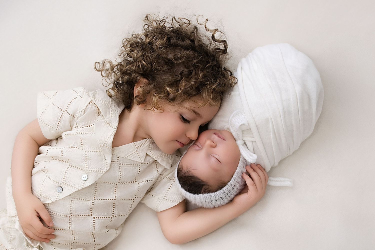 Two siblings in soft neutral outfits lying down together in a gentle embrace.
