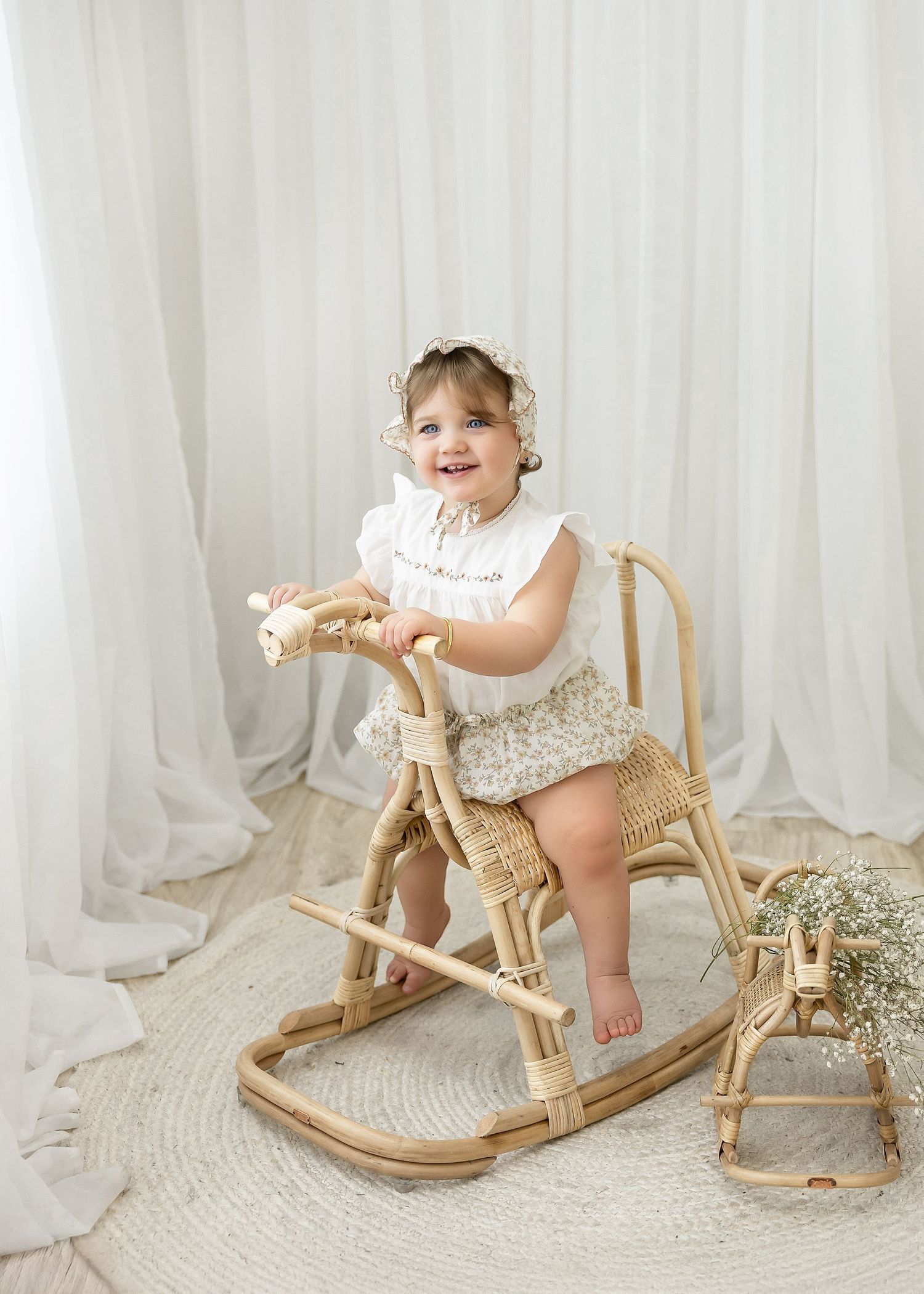 A small infant wearing white plays with a wooden rocking horse on a beige rug by white curtains.