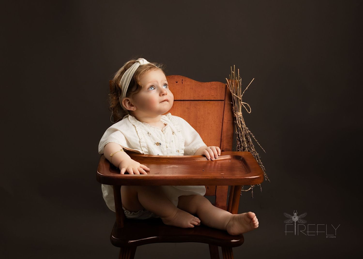 A small child wearing vintage-style clothing sits in an antique wooden high chair against a dark background with dried wheat stalks.