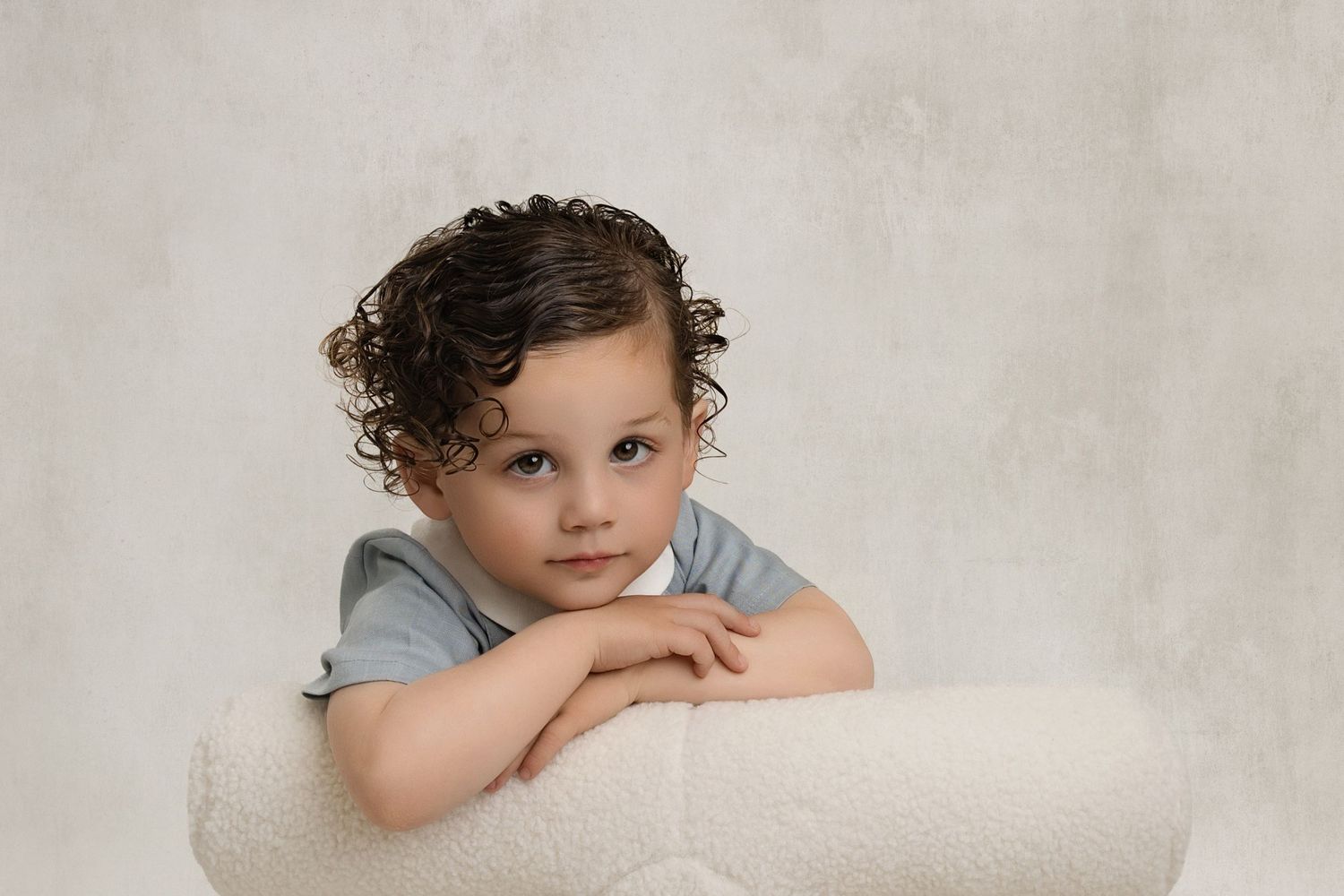 Studio portrait of a child in grey outfit leaning on white cushion.