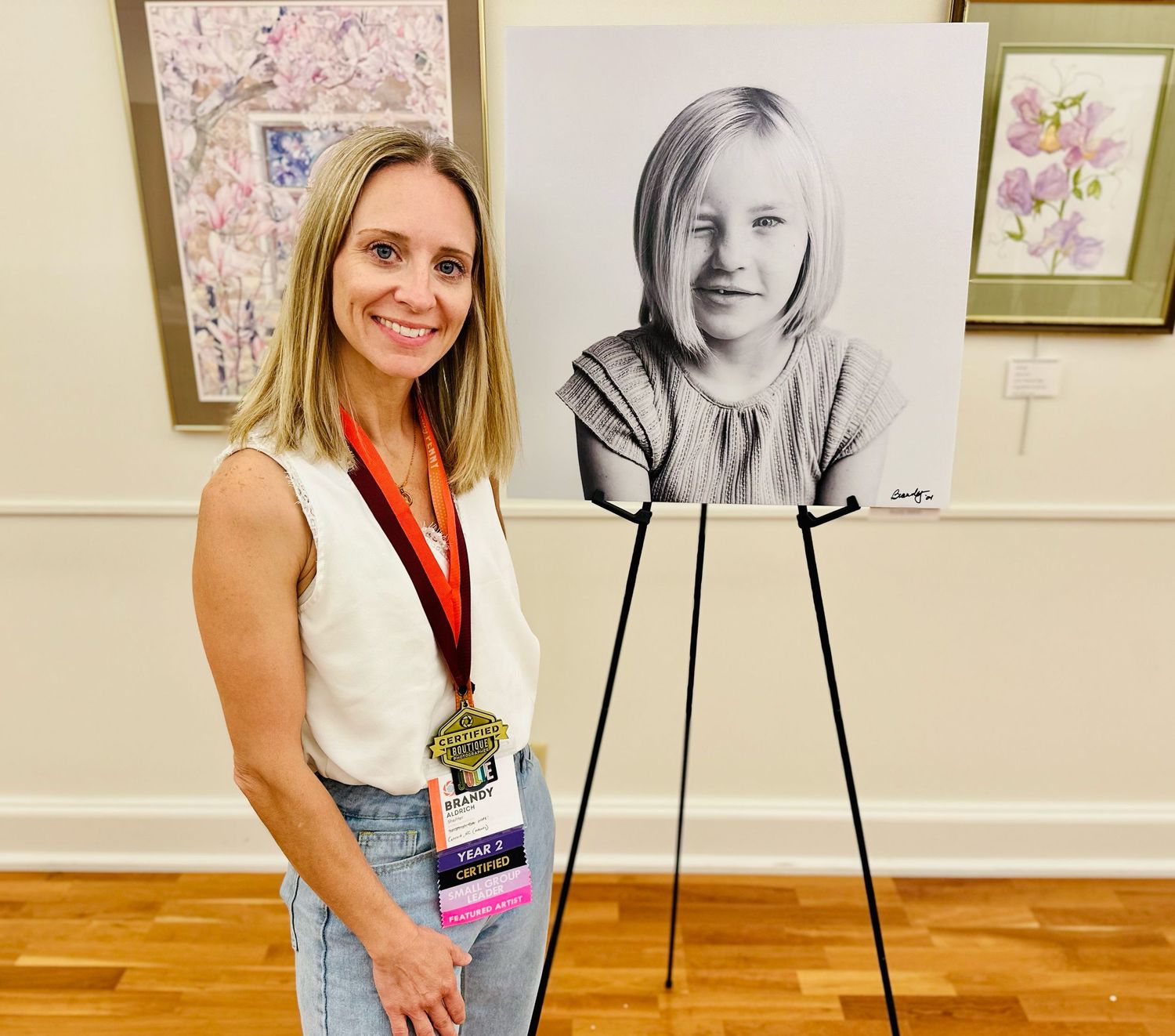 Person posing next to charcoal portrait drawing displayed on easel in wooden floored gallery.