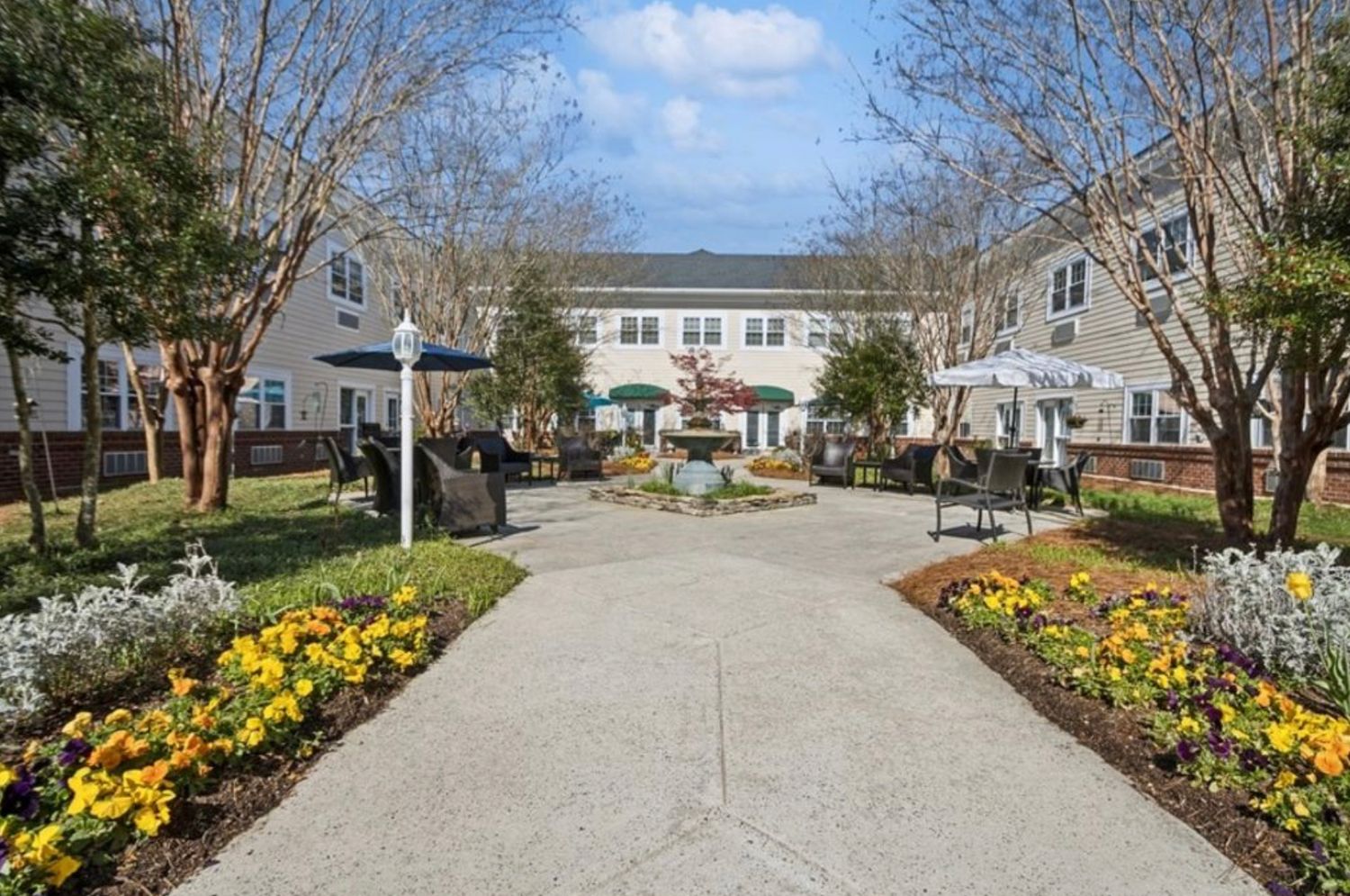Scenic courtyard pathway lined with colorful yellow and purple flowers leading to a charming multi-story building with outdoor seating.