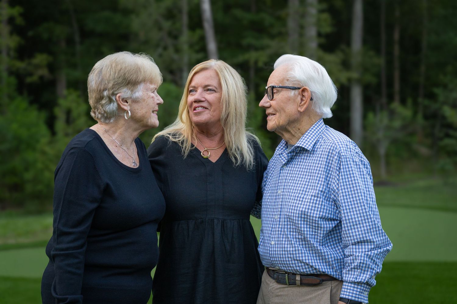 Three people smiling and conversing outdoors on a green lawn with trees in the background.