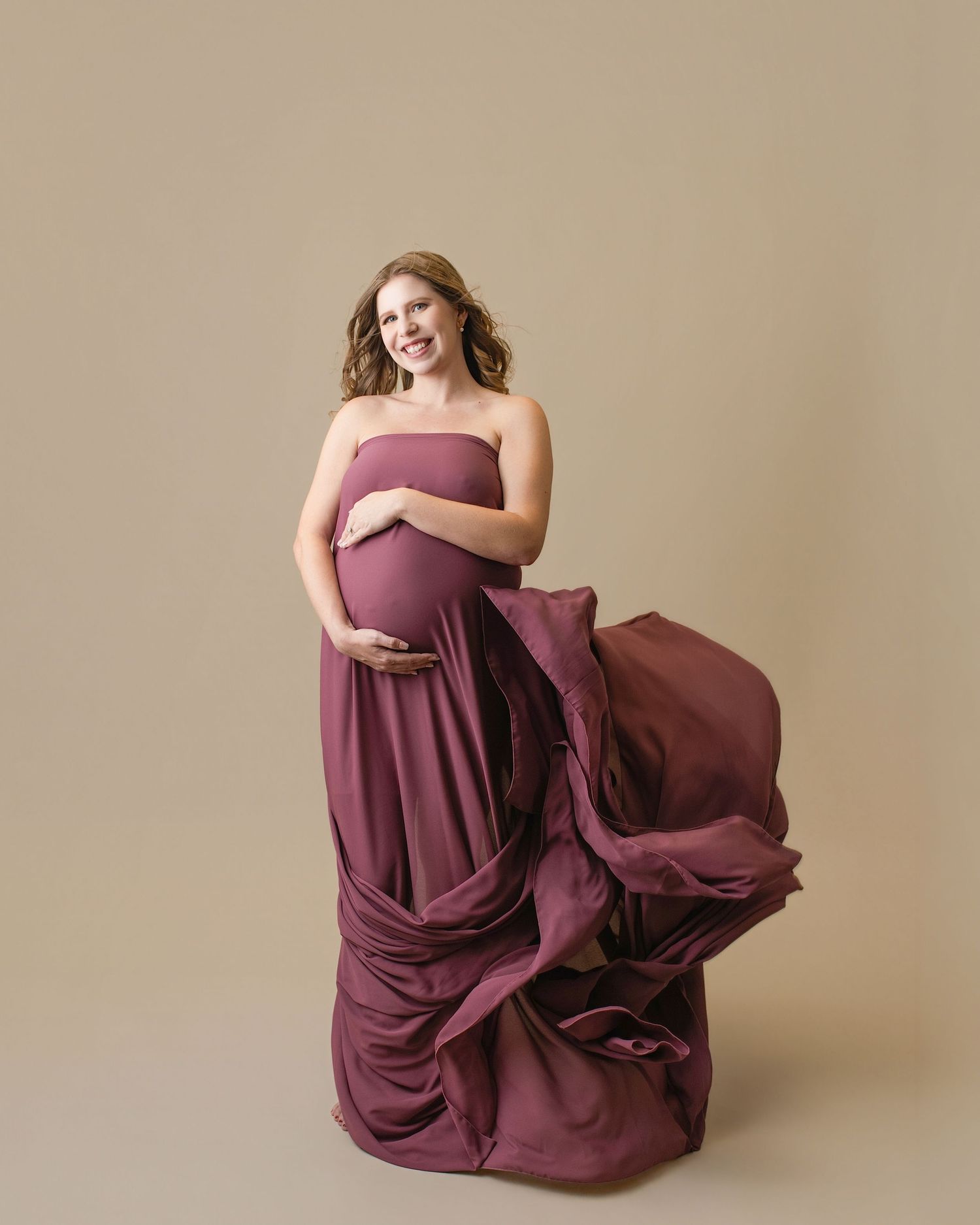 Pregnant woman in flowing mauve strapless gown cradling baby bump, fabric billowing gracefully against neutral studio background.
