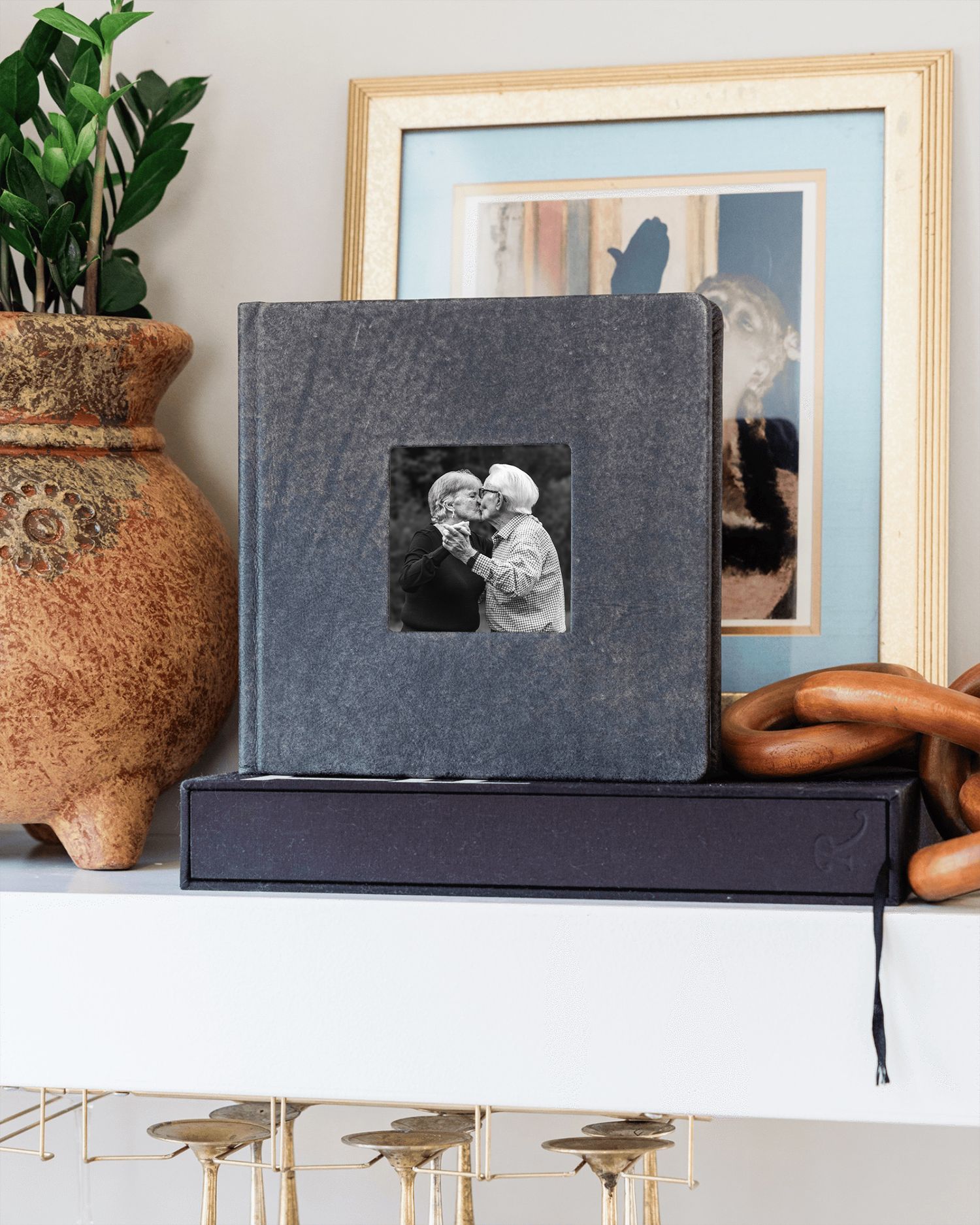 Gray linen photo album with black-and-white couple photo on mantel beside terracotta vase and framed artwork.