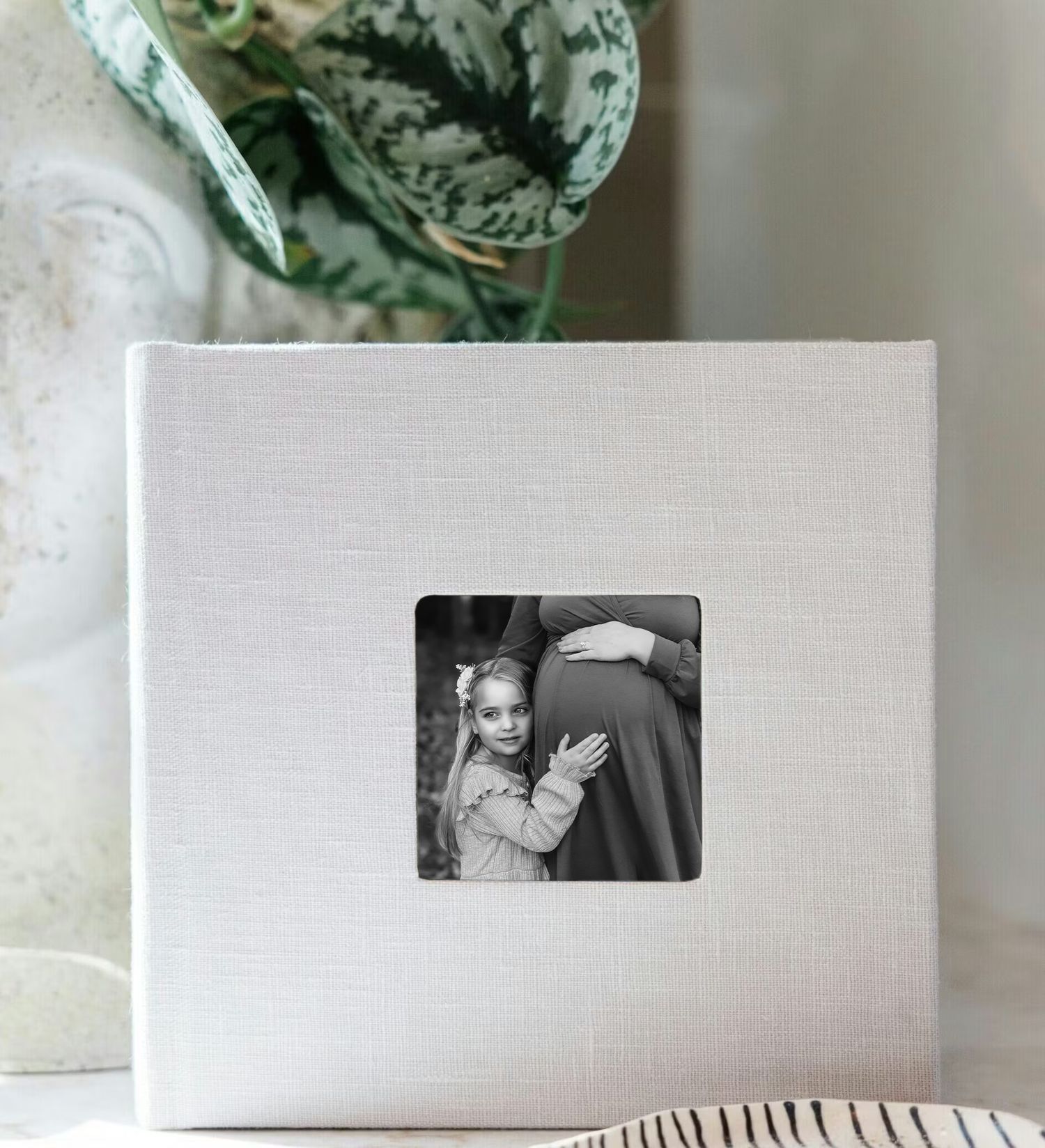 White linen photo album with a small square window displaying a black and white maternity photo of a girl hugging a pregnant woman.