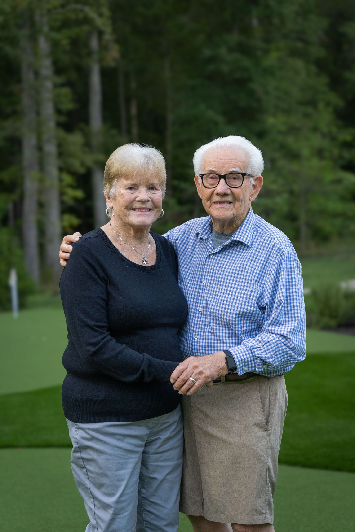 Smiling senior couple embracing outdoors on a lush green golf course surrounded by tall trees.