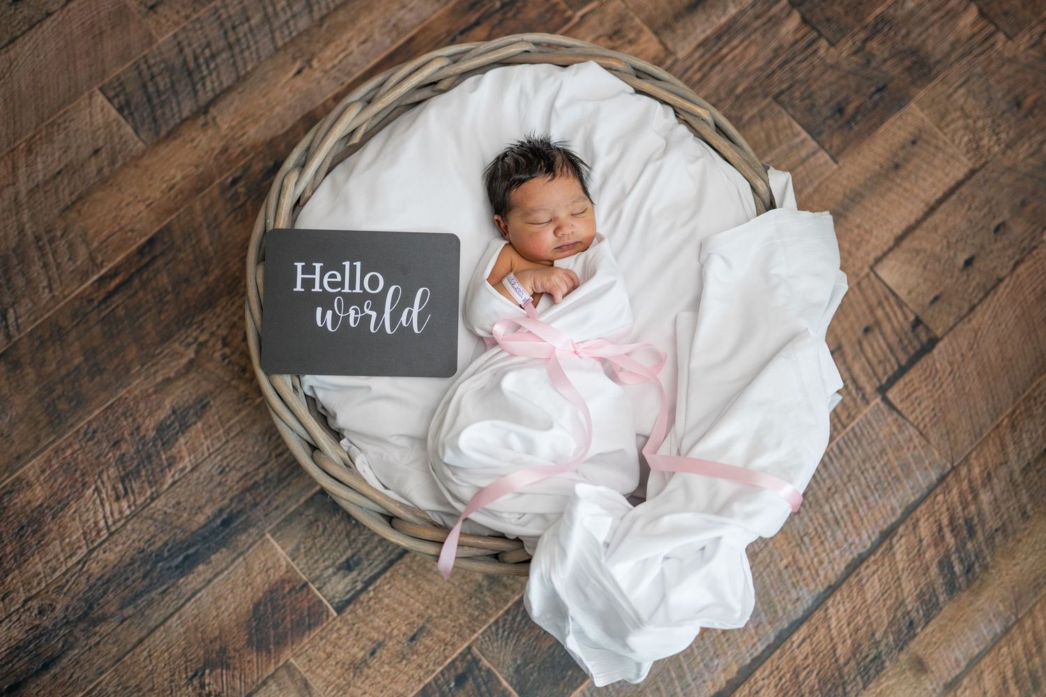 Newborn baby girl portrait. Baby is swaddled in white with a pink bow, and posed asleep in a basket. Next to her is a si