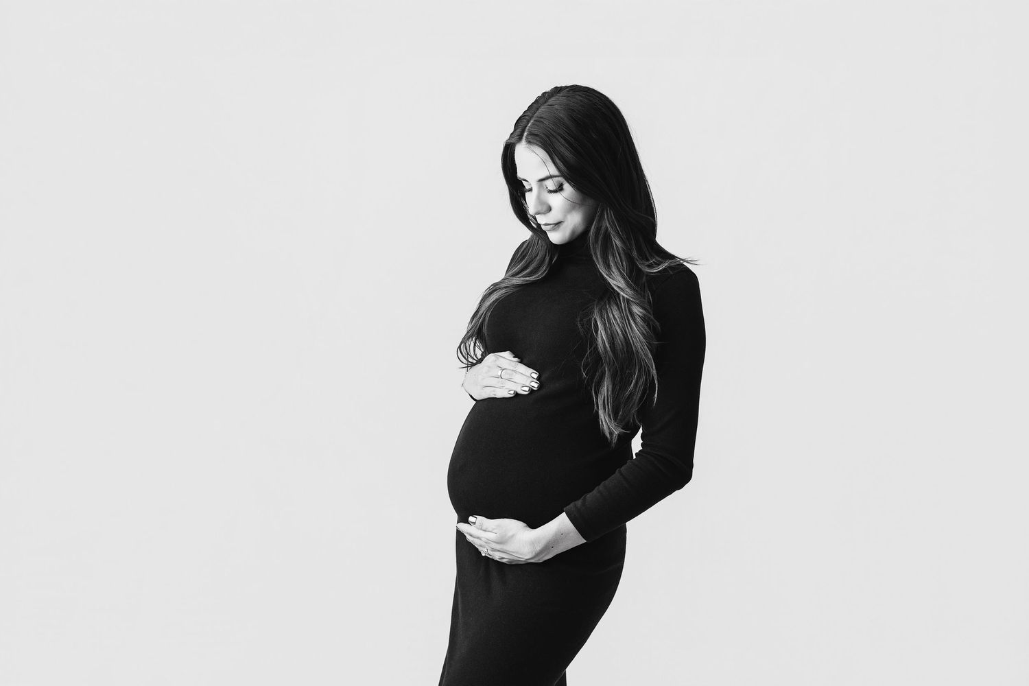 Black and white photo of a pregnant woman in a black dress cradling her baby bump against a white background.