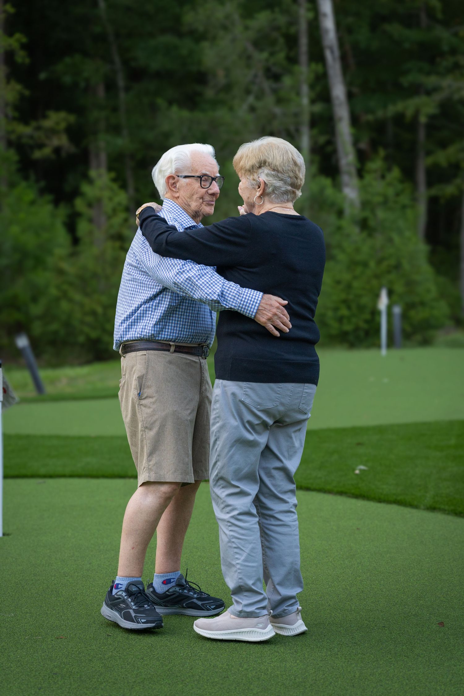 Two elderly people embracing warmly on a golf course green, surrounded by lush trees in the background.