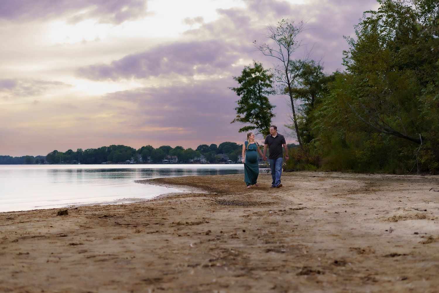 Couple walking hand in hand on sandy beach at sunset with calm water and lush green trees in background.