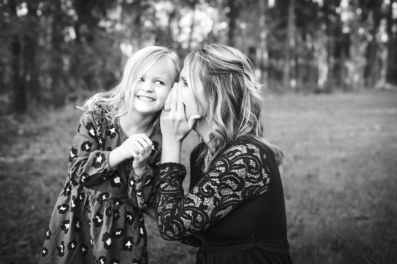 Black and white photo of a mother whispering to her smiling young daughter outdoors in a wooded setting.