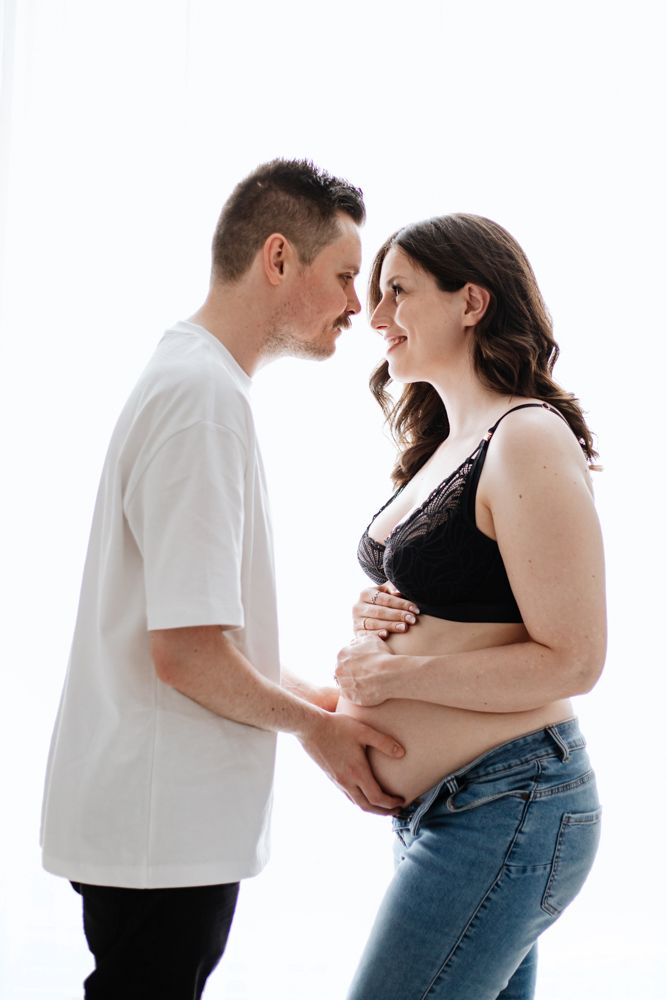 A couple shares intimate moments in their maternity photoshoot against a white background.