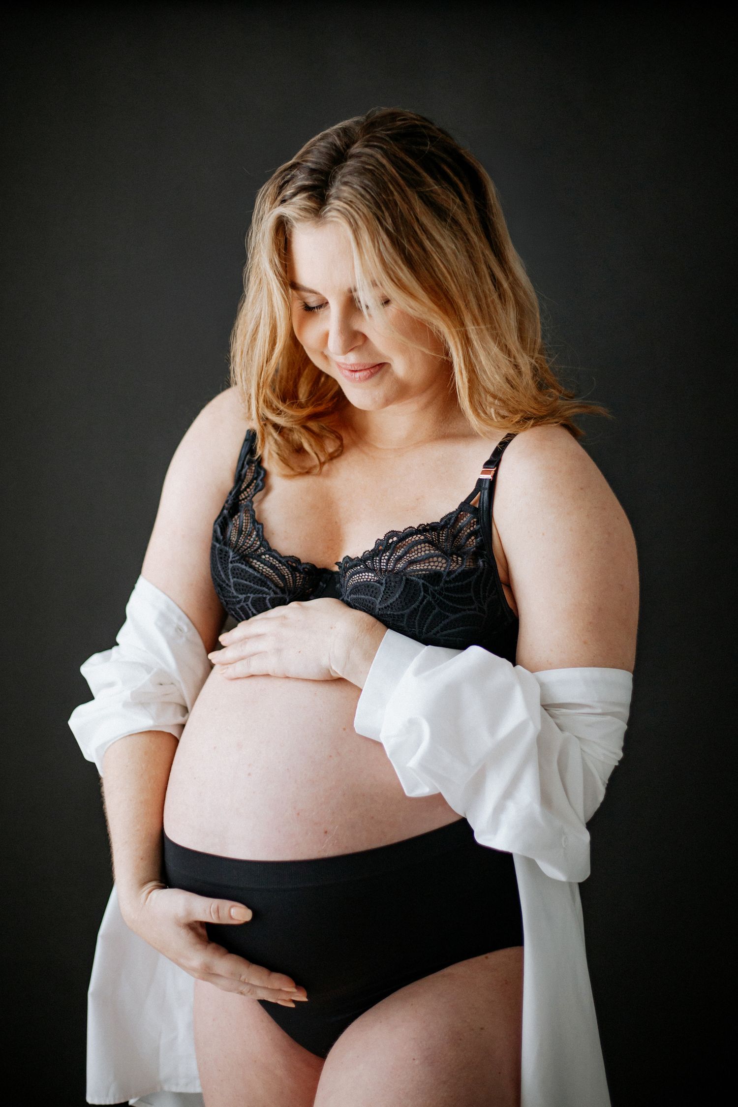 Pregnant woman in black lingerie and white cardigan poses against dark background during maternity photoshoot.