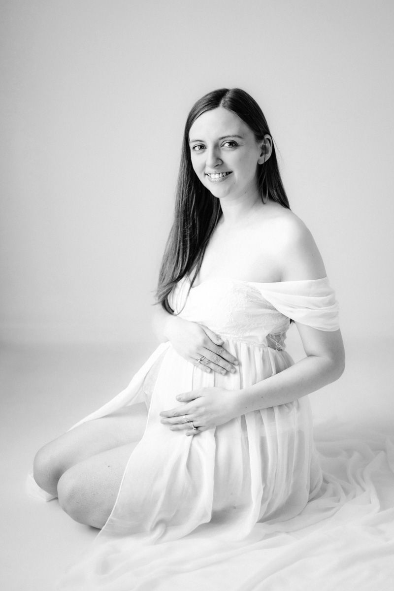 Black and white maternity photo of a smiling pregnant woman in white draped white dress, sitting on her kneews cradling her baby bump.