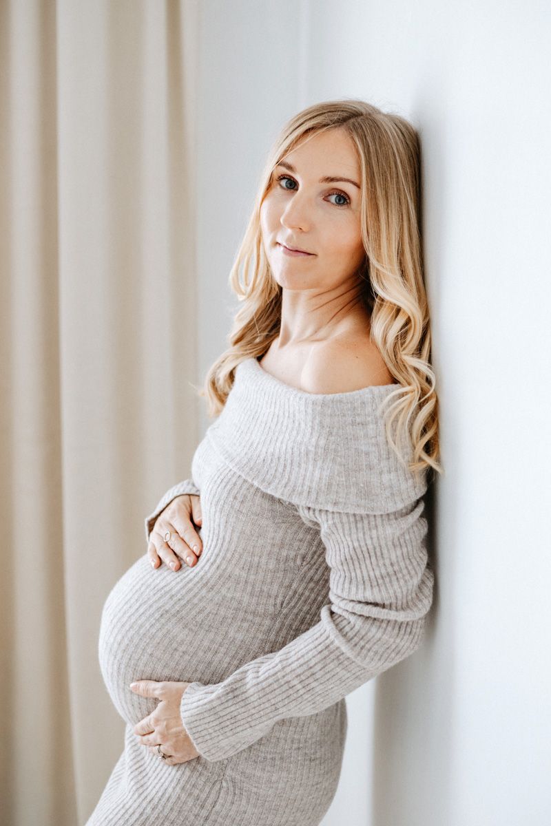 A pregnant woman in a cosy gray sweater dress poses against a light background in a maternity photo series.