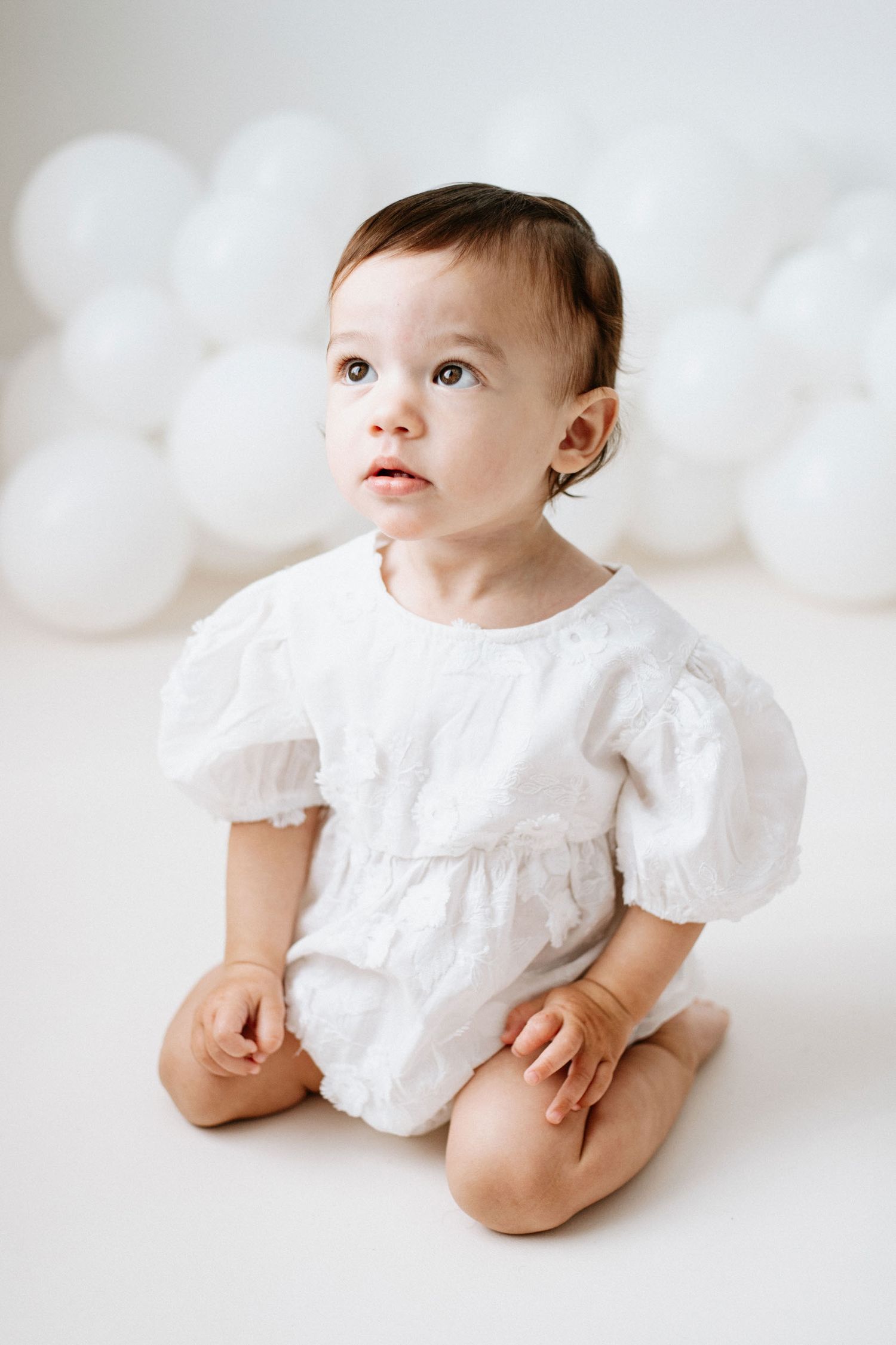 A baby in a white dress lies on a soft white backdrop during a professional photo shoot.