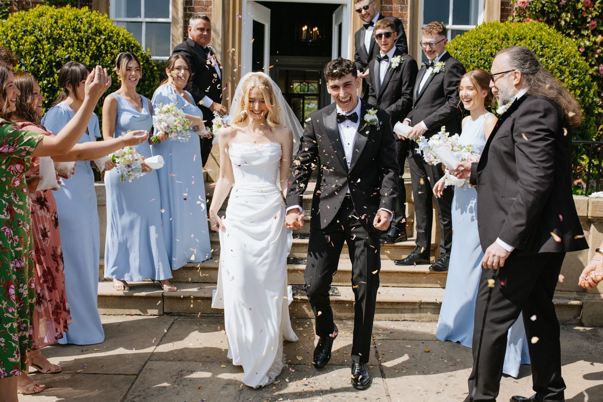 Bride and groom walk through confetti shower with wedding party in blue dresses outside Norwood Park Wedding venue in Nottinghamshire