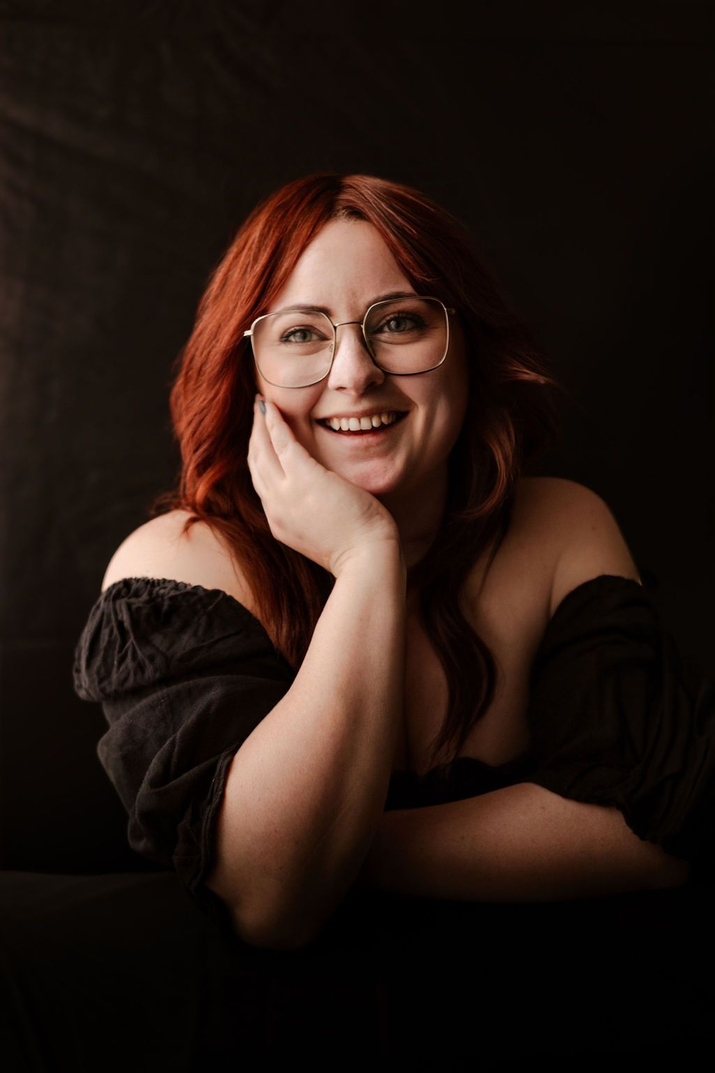 Portrait of someone in a black off-shoulder top and glasses against dark background with warm lighting and natural smile.