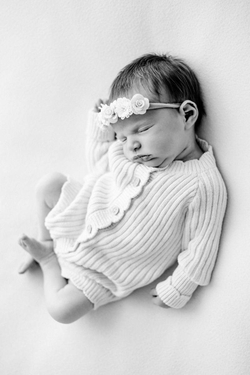 Black and white photo of a sleeping newborn baby girl wearing a knit romper and floral headband.