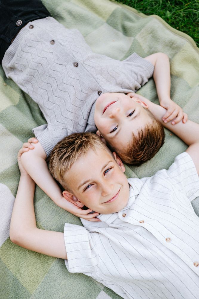 Two children lying side by side on a blanket outdoors, wearing light colored shirts and smiling at the camera.