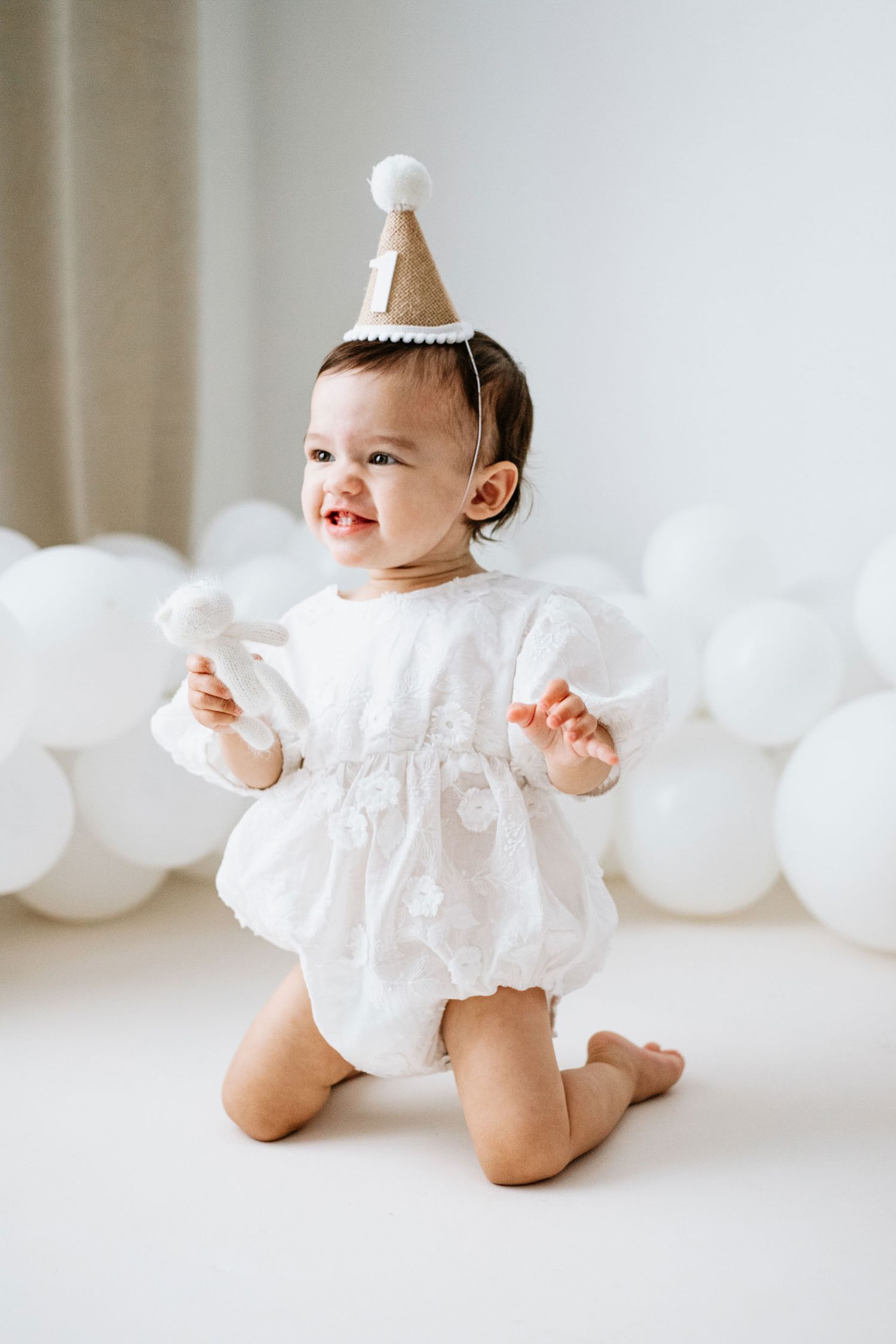 Baby wearing white dress and party hat sits on white floor with balloon decorations in background.