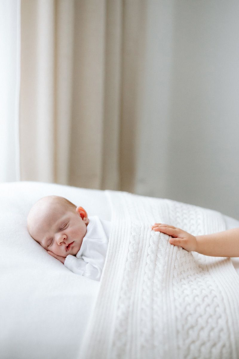 Newborn baby sleeping peacefully on white bed while sibling hand gently touches knit blanket draped over the baby.